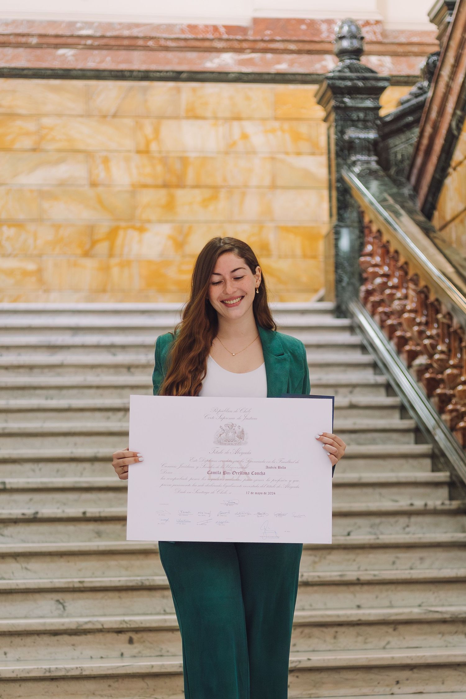 Happy young woman in emerald green suit displaying official diploma on grand staircase.