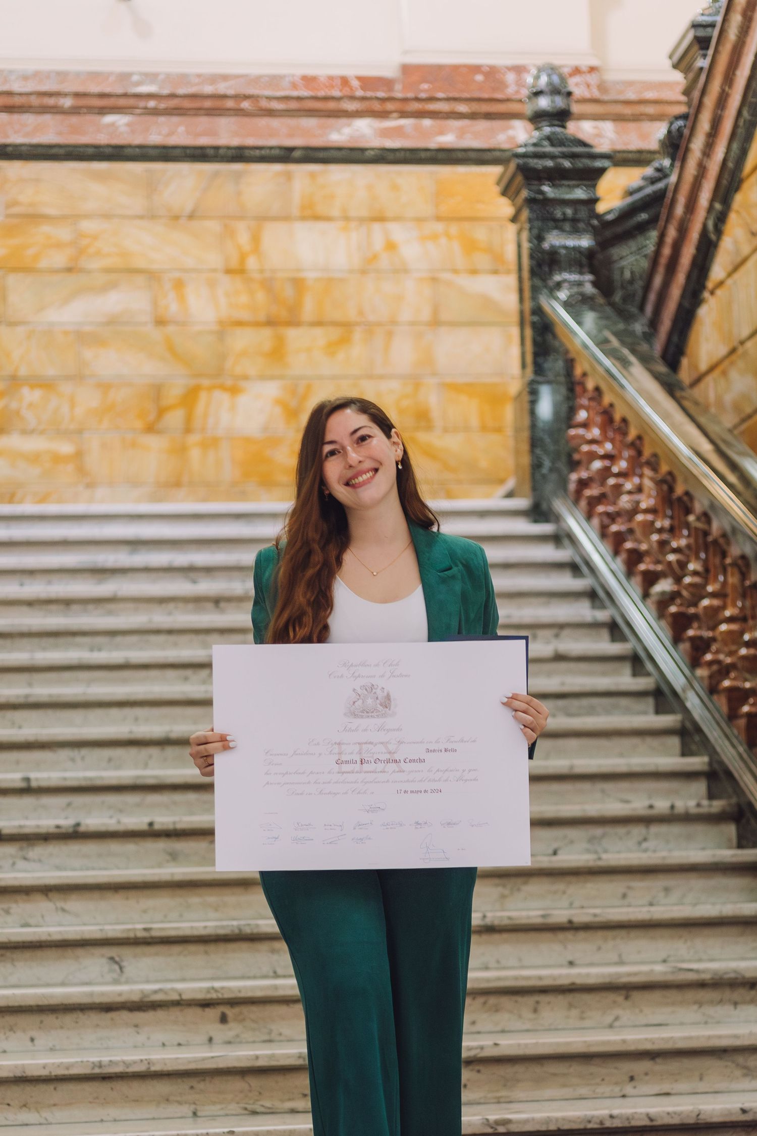 Woman in green pantsuit proudly holding large white certificate on ornate marble stairs.