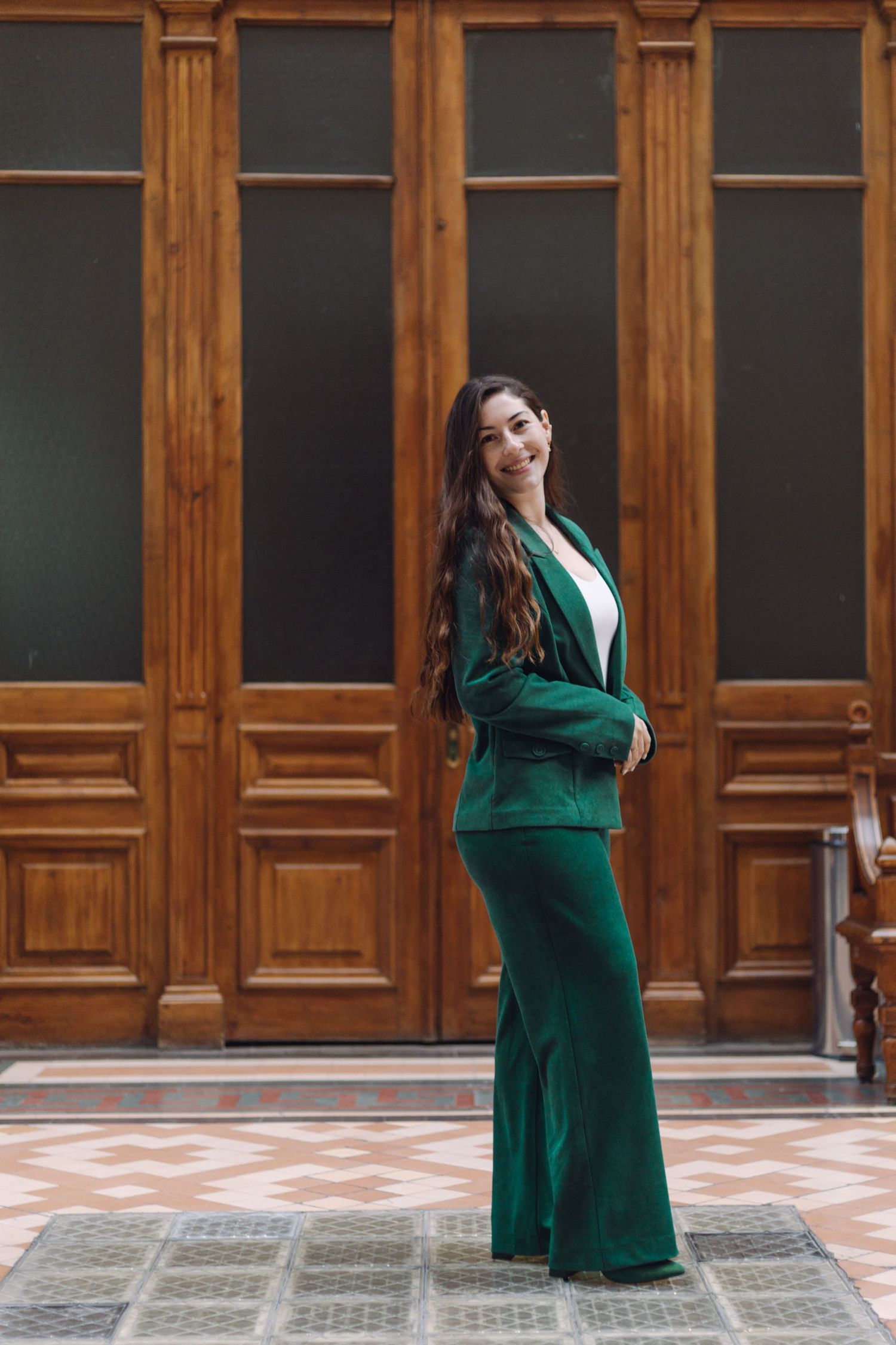 Elegant woman in emerald green suit posing before classic wooden doors on tiled floor.
