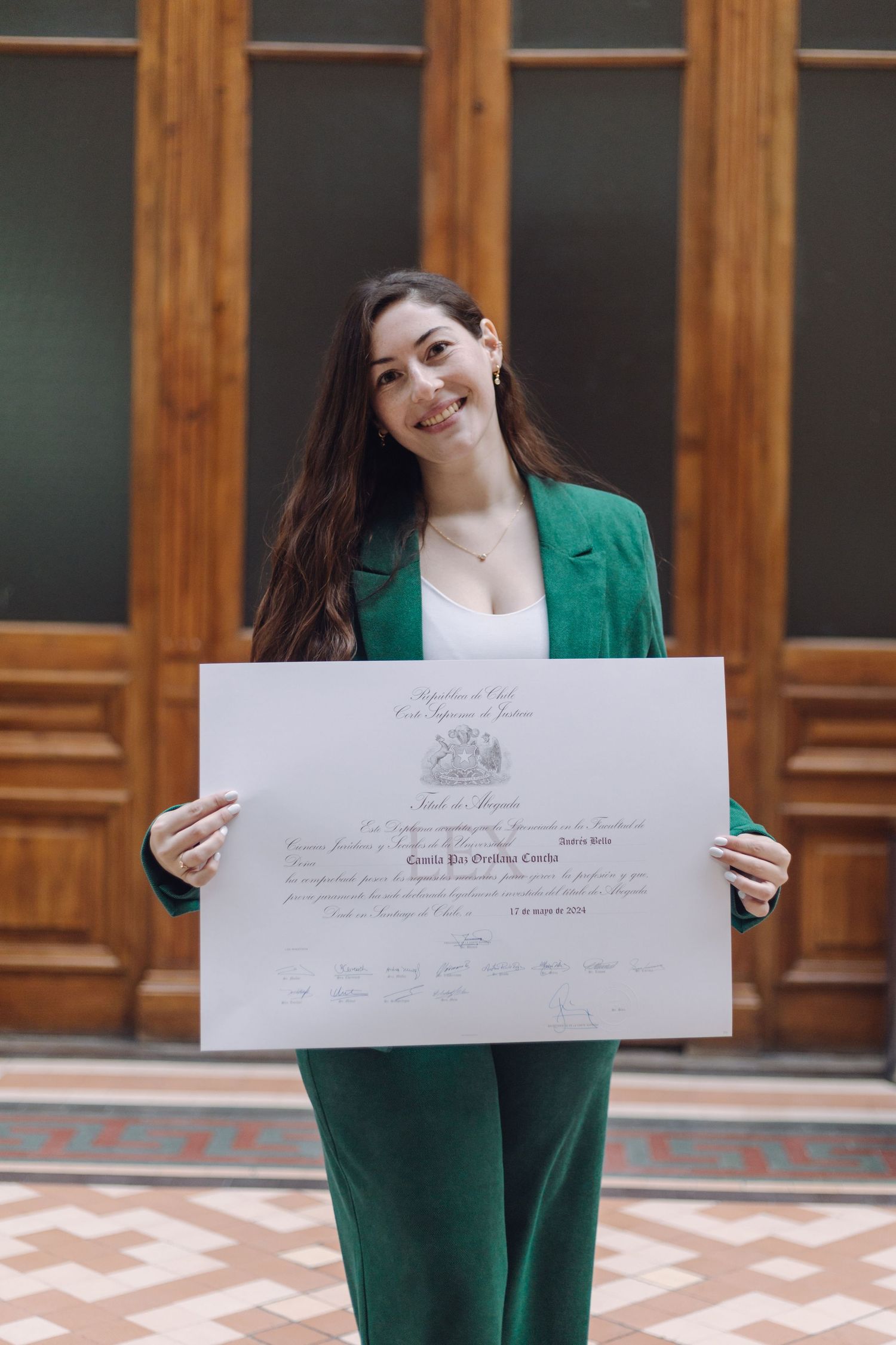 Smiling graduate in green suit displaying large official Chilean judiciary certificate outdoors.