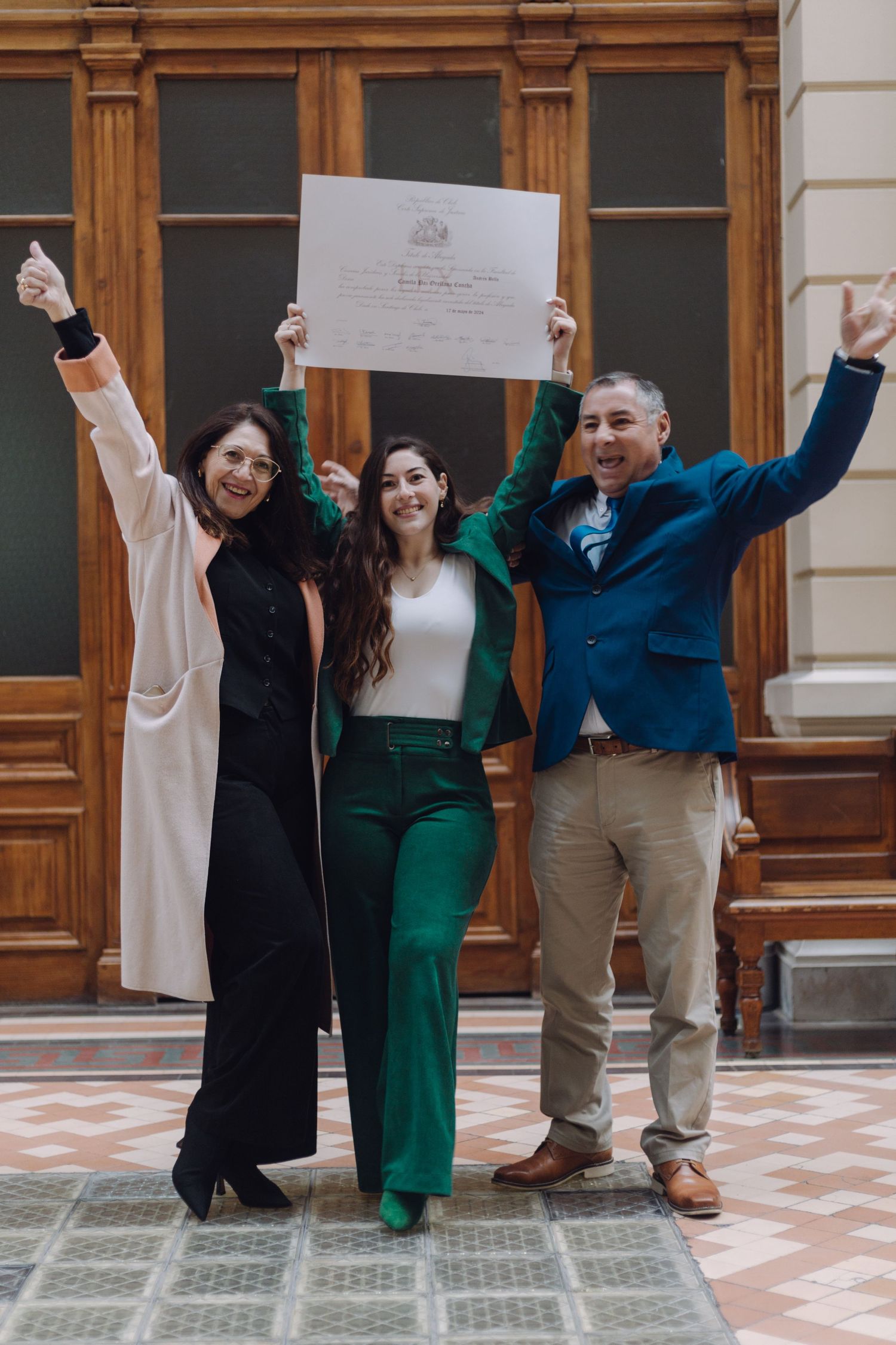 Three people celebrate a graduation holding diploma aloft with arms raised in a classic building.