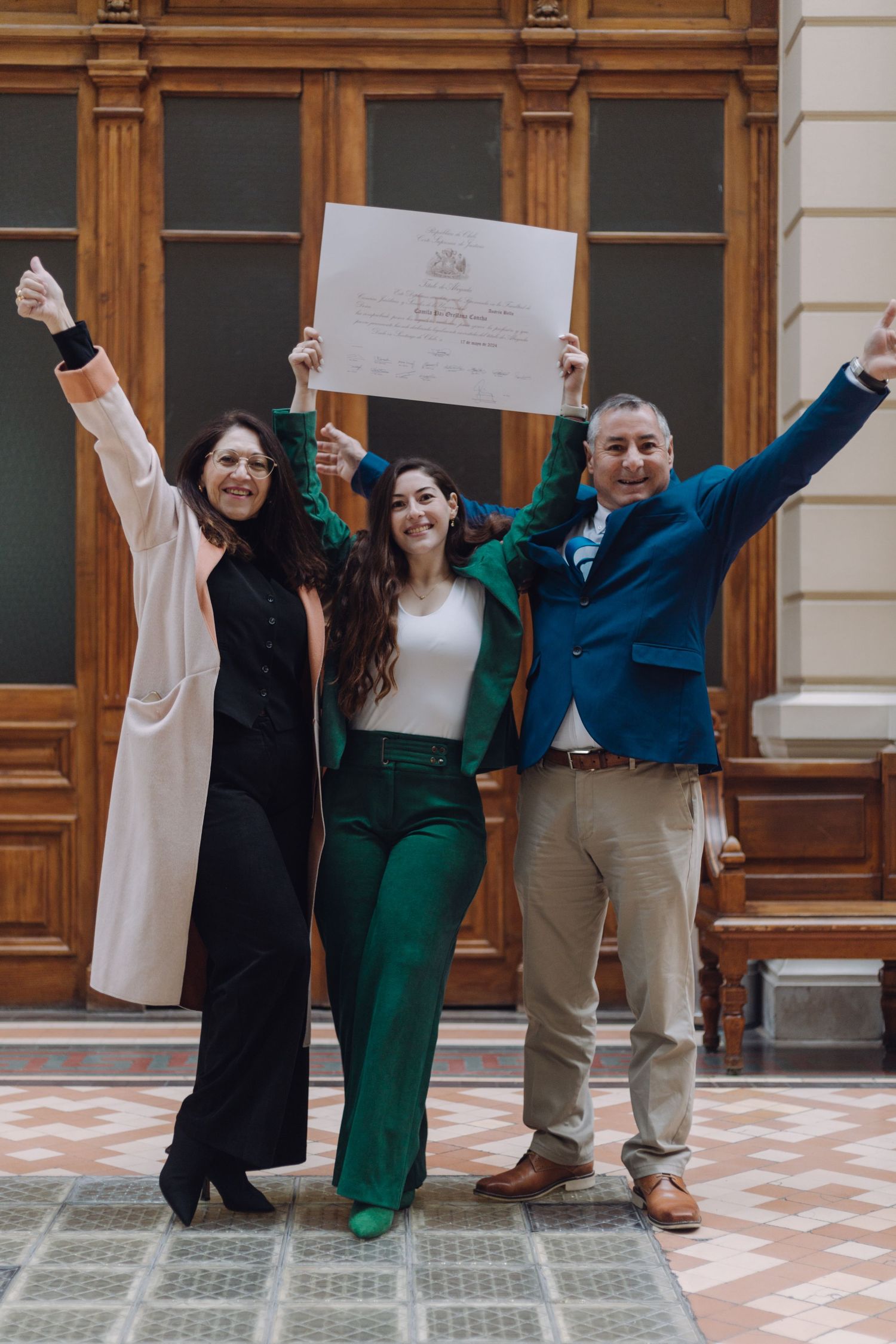 Joyful family trio raises arms celebrating graduation with diploma in ornate building.