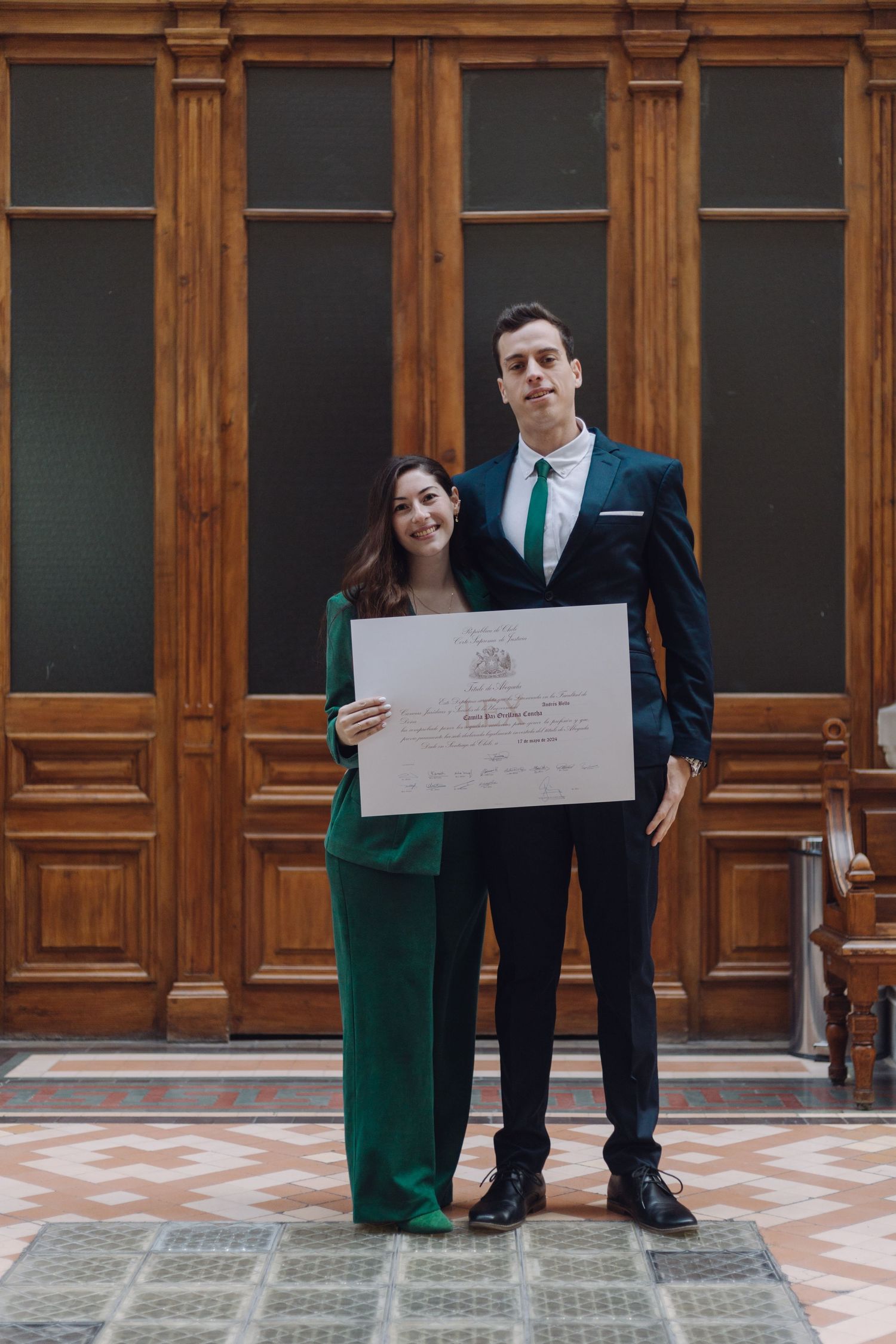 Graduate in green suit holds diploma with suited companion before wooden doors.