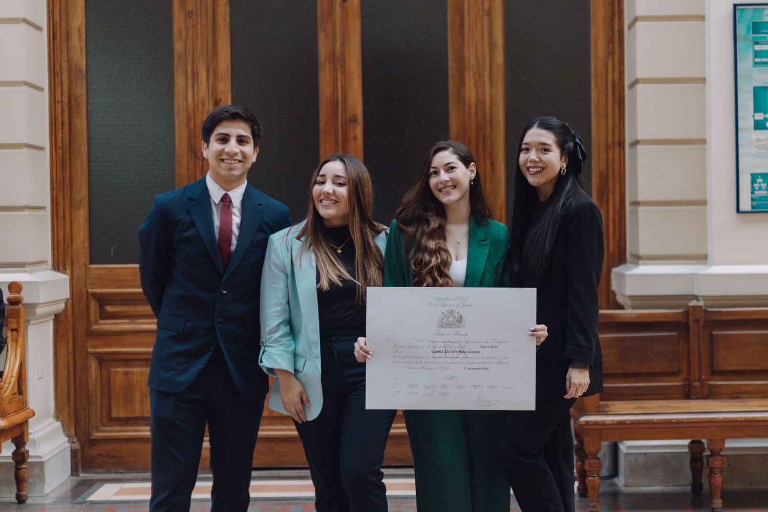 Four smiling graduates display diploma together in a formal academic setting.