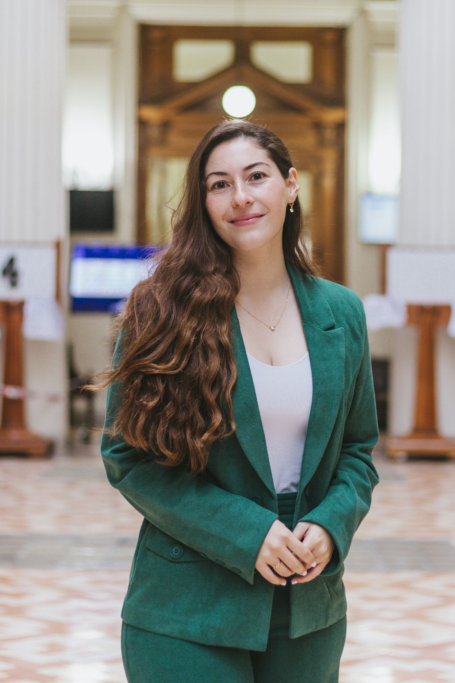 Young woman in green suit smiles confidently in grand columned hall interior.