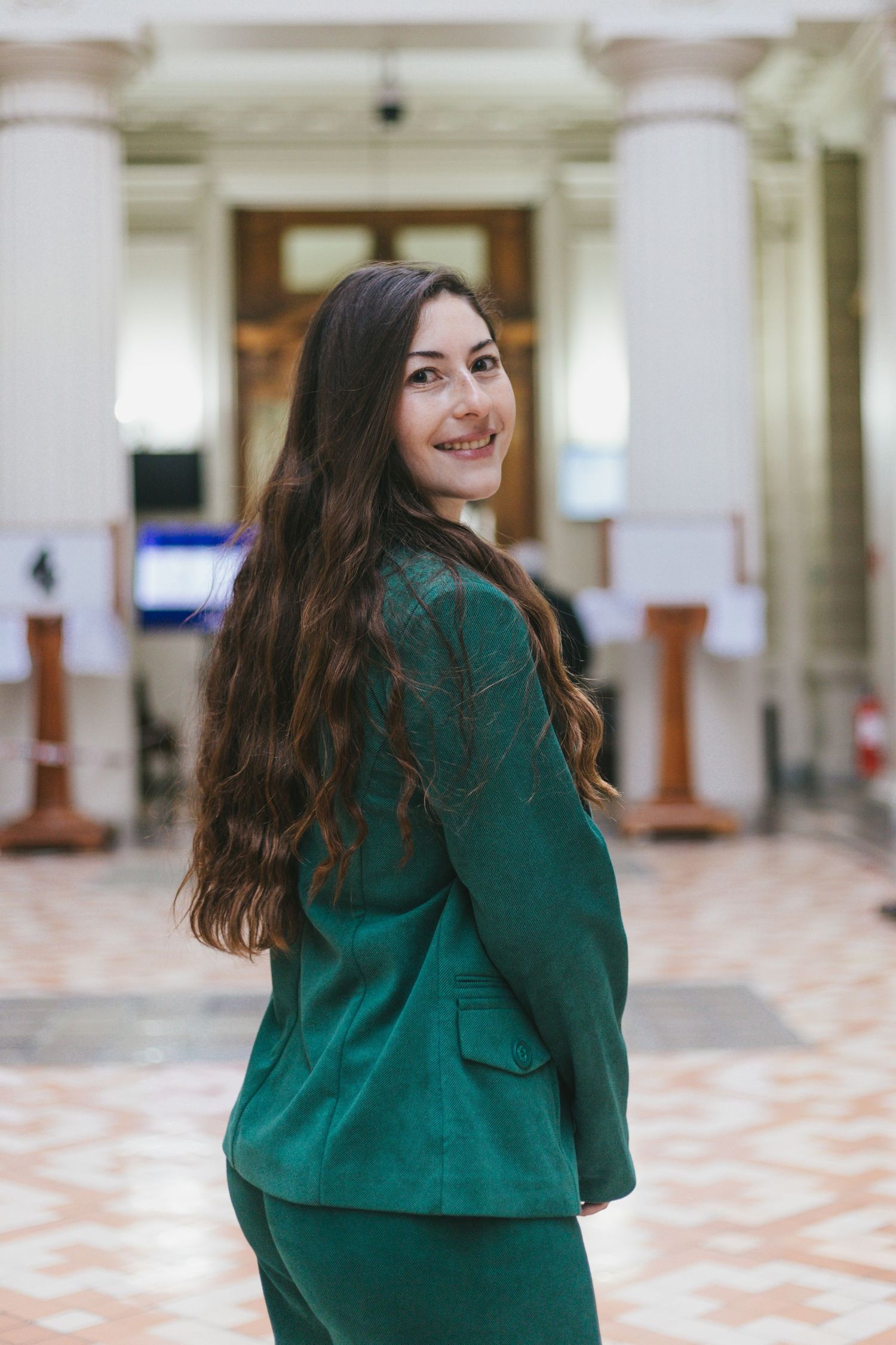 Cheerful young woman in green suit glances back smiling in grand hall.