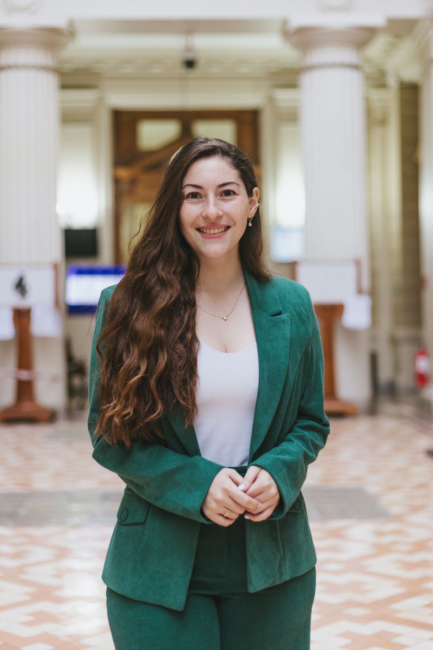 Smiling young woman in elegant green suit stands in bright columned hall.