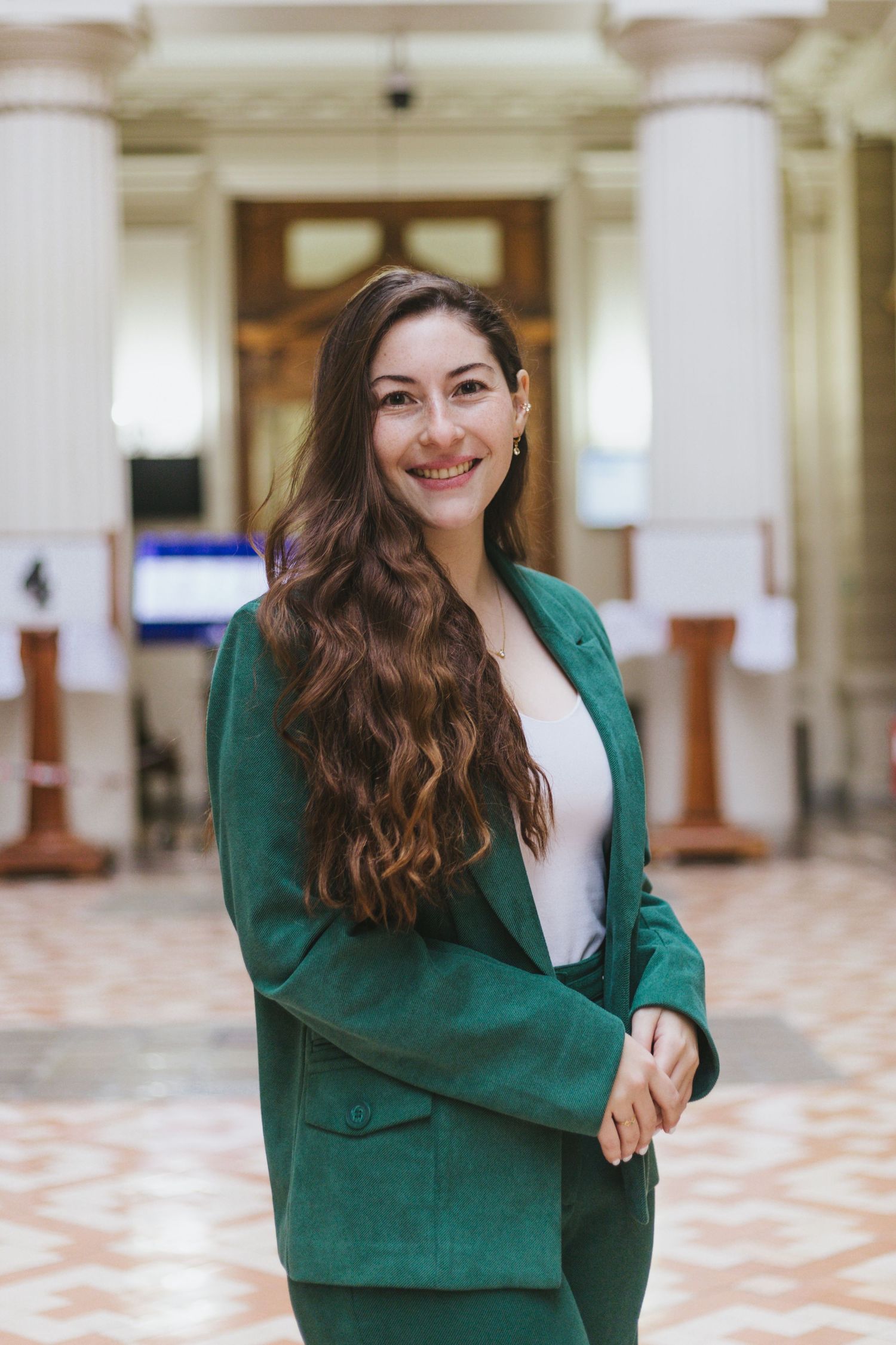 Confident young woman in green blazer smiles in bright columned building interior.