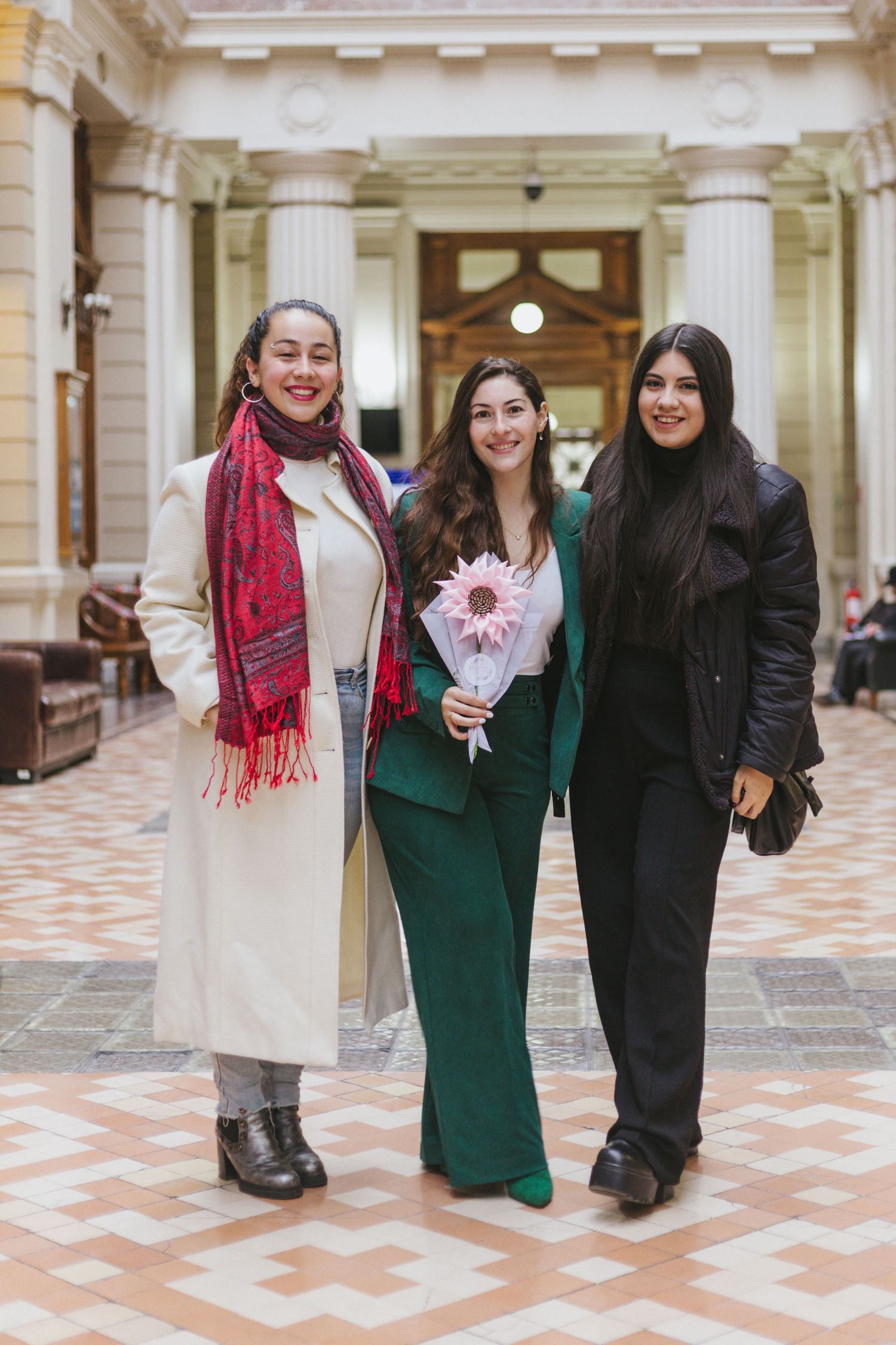 Three smiling women walk together in a grand interior hall, one wearing green suit holding a pink flower bouquet.