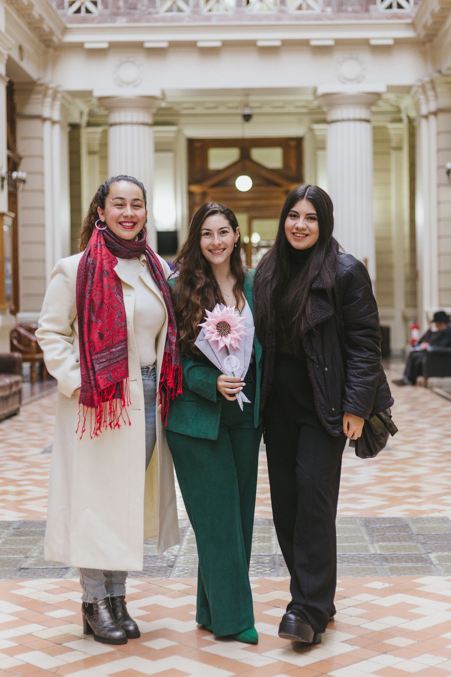 Three women pose together indoors in an elegant hall, one in green suit holding pink flower, another in white coat with red scarf.