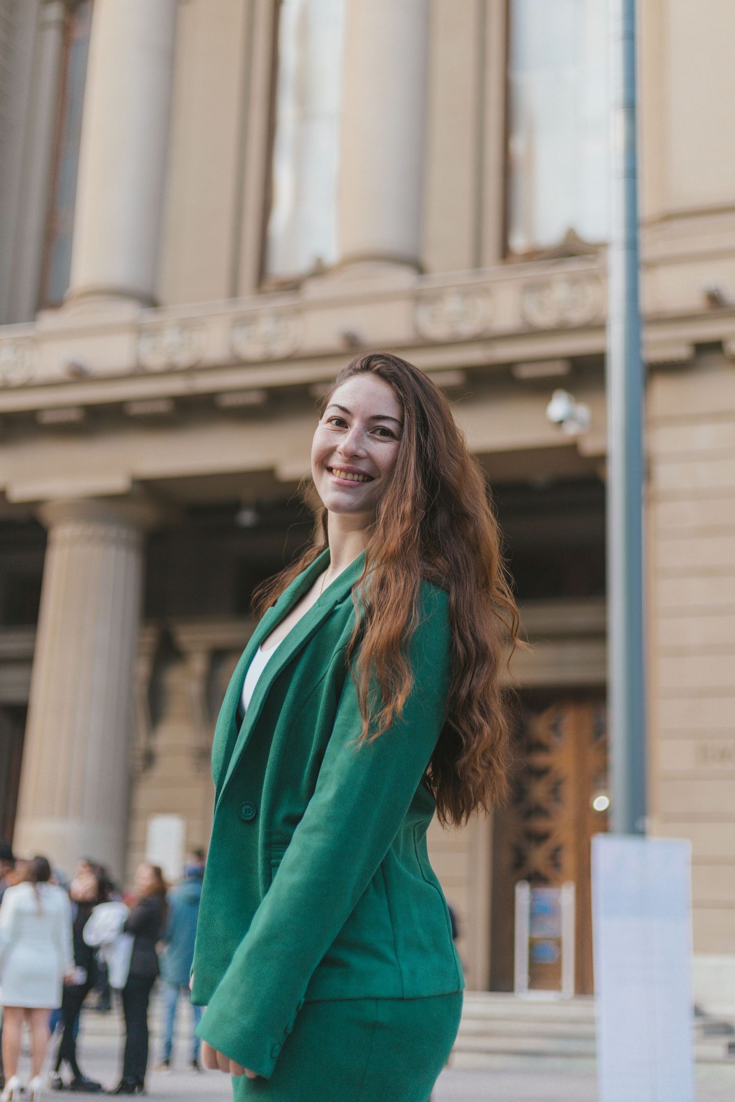 Smiling woman with long hair wearing a green suit poses outdoors in front of a classical stone building with columns.