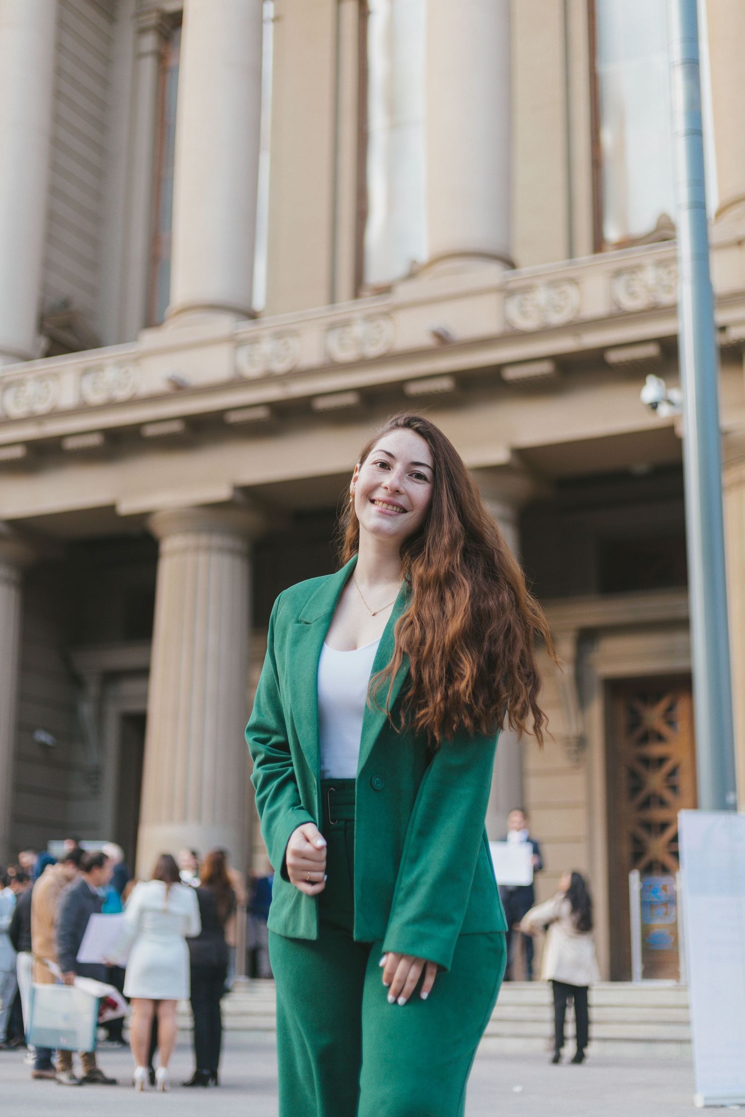 Young woman in green blazer and pants smiling outdoors in front of a neoclassical building with tall columns.