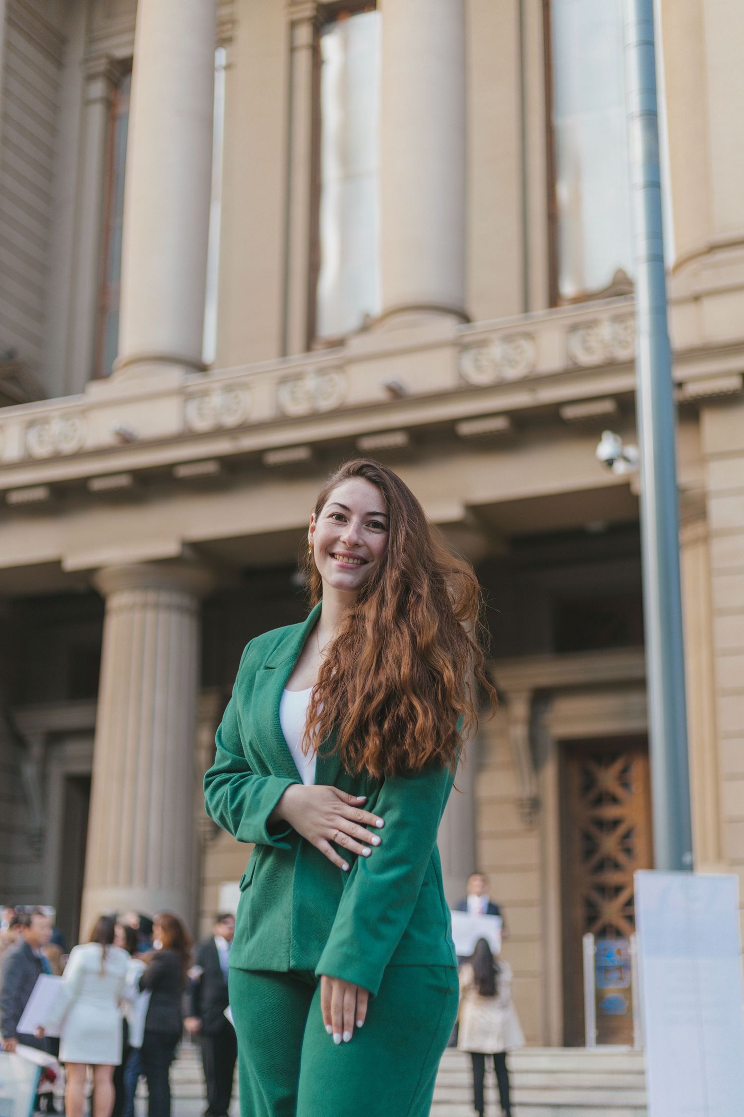 Elegant woman in green suit smiling confidently outside a neoclassical building with large columns in background.