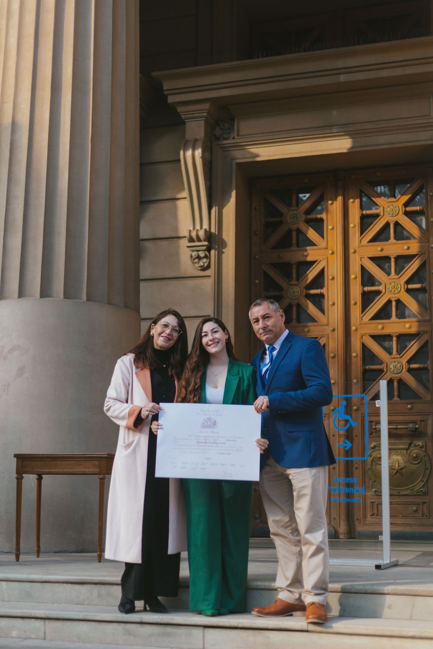 Three people proudly display a graduation certificate on steps of a grand building with ornate wooden doors.