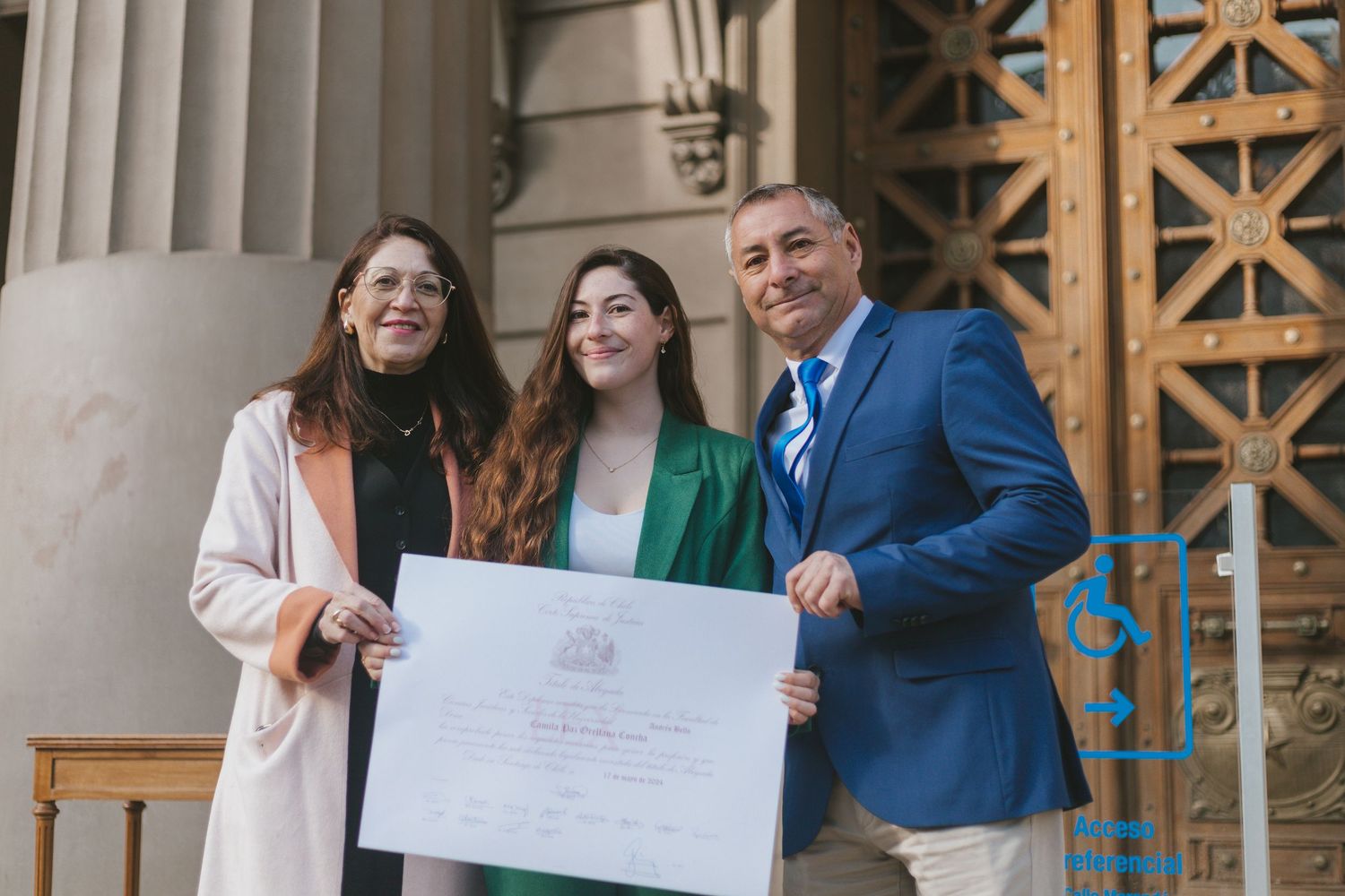 Smiling graduate in green suit poses with family holding a large diploma certificate outside a formal building.