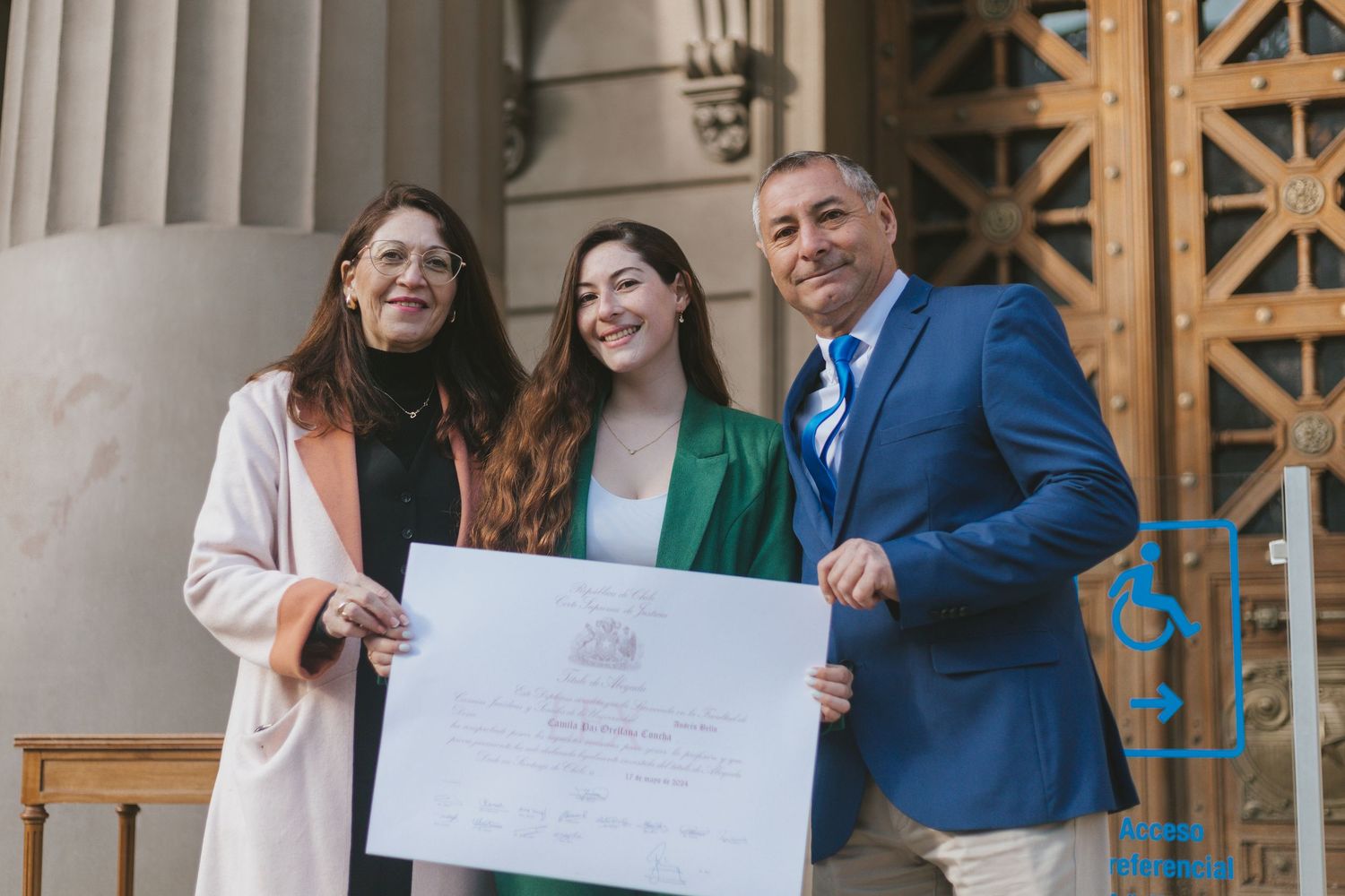 Graduate in green suit holds diploma certificate with two proud family members outside a classical building.