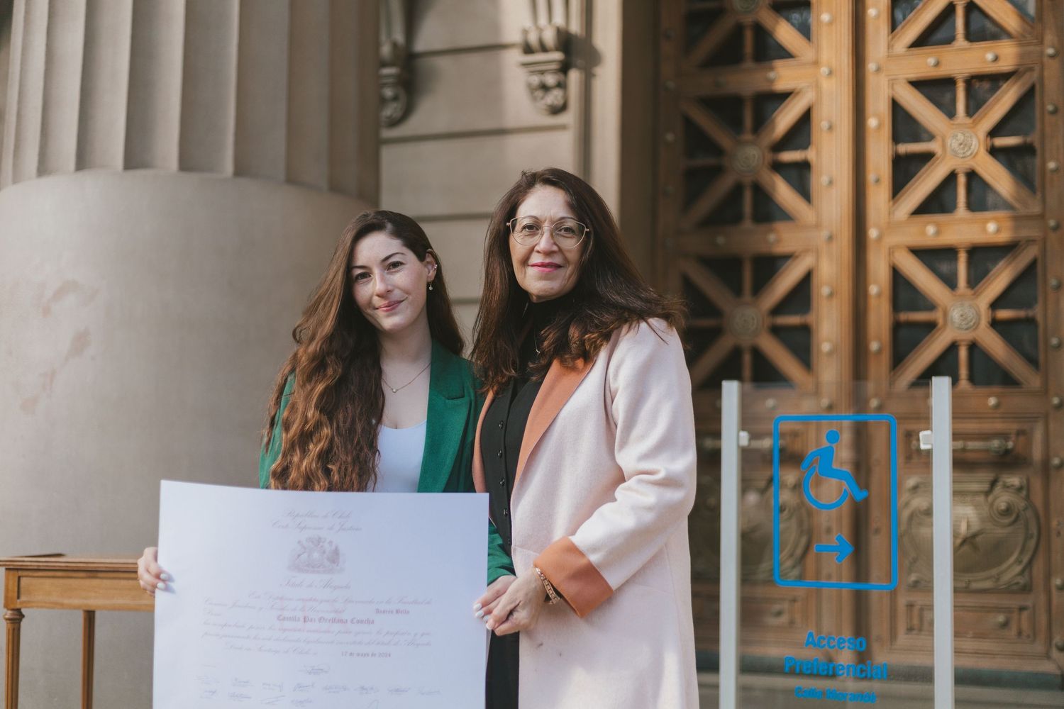 Graduate in green suit and older woman in pink coat proudly display a large diploma certificate outdoors.