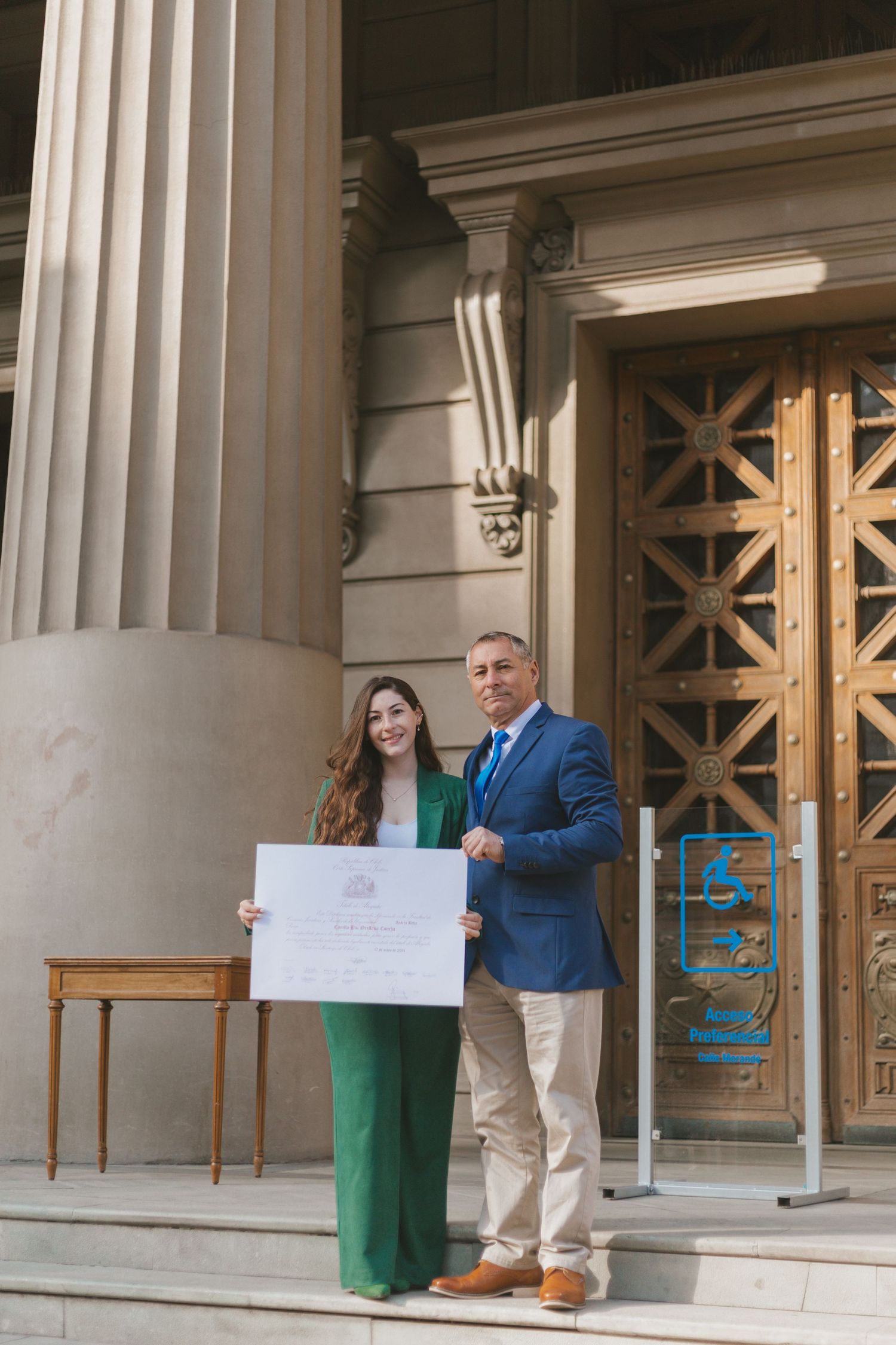 Graduate in green suit and man in blue blazer hold a large certificate on steps of a neoclassical building.