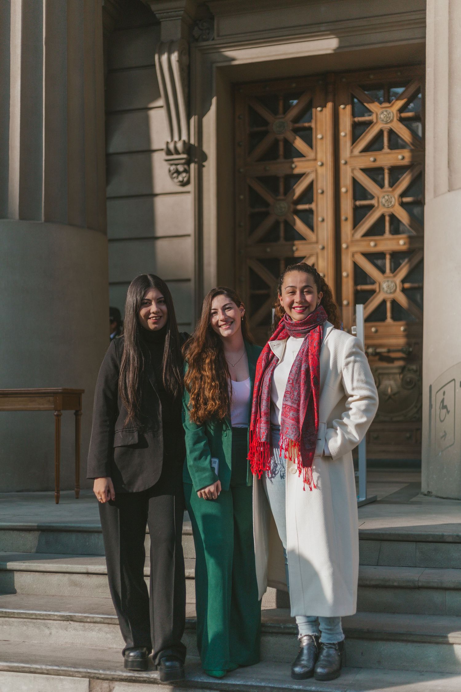 Three women smiling on building steps, wearing stylish outfits including green suit, black coat, and white coat with red scarf.