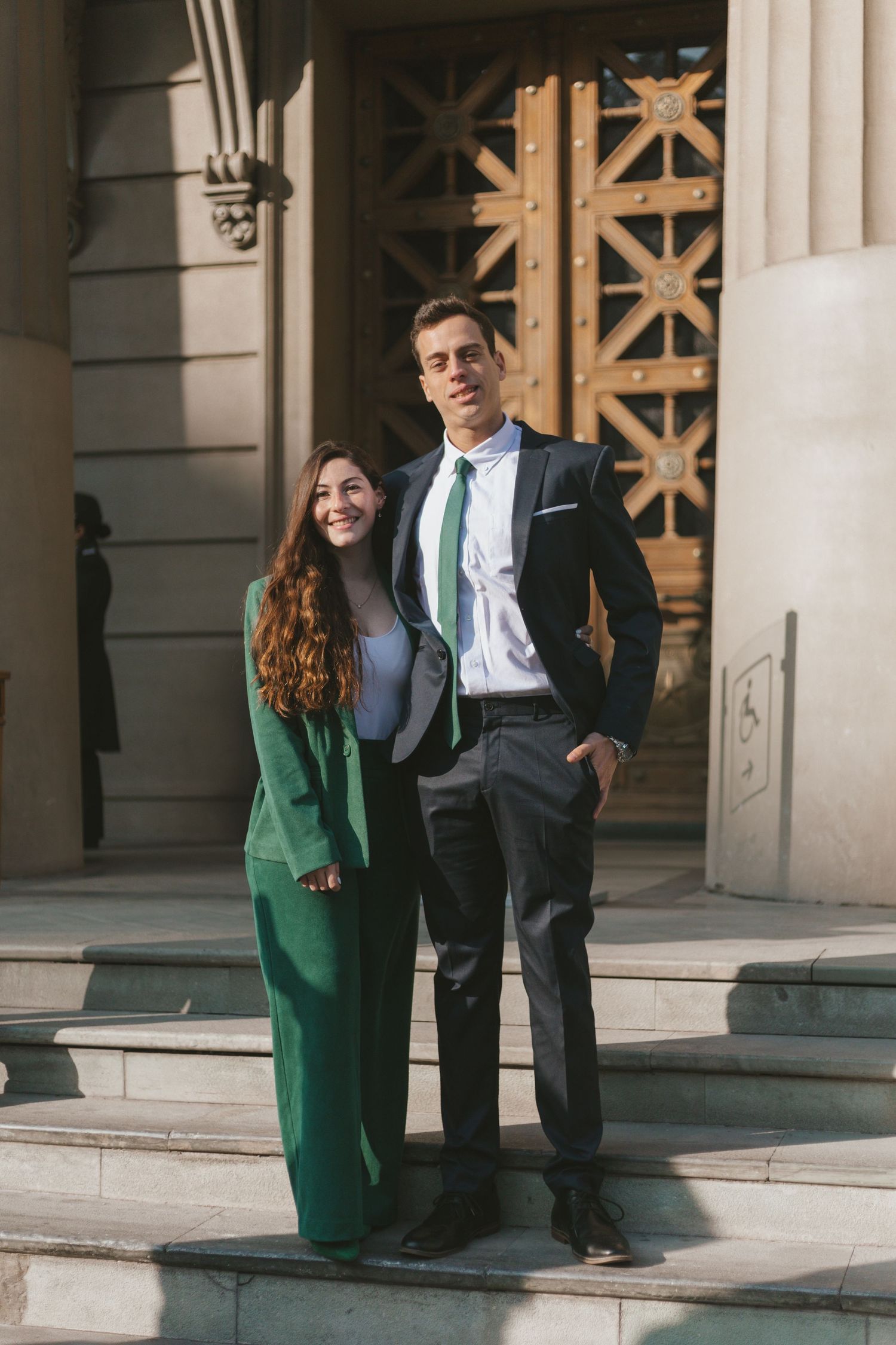 Couple in formal attire posing on steps of a classical building, woman in green suit and man in dark suit with green tie.