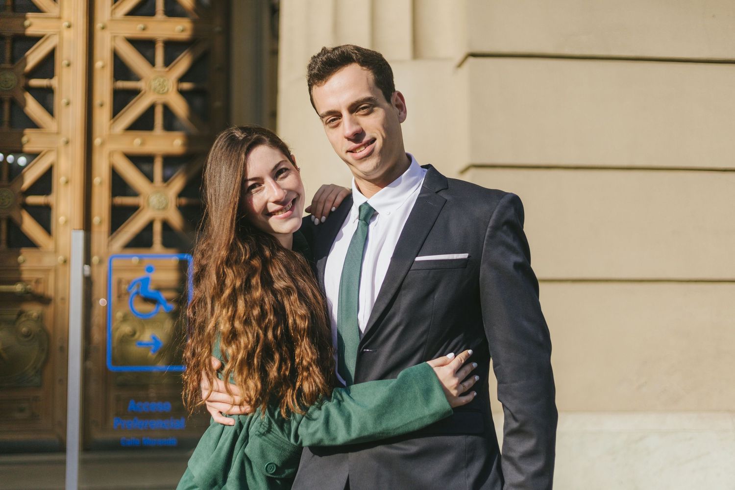 Smiling couple embracing outdoors, woman in green blazer and man in dark suit with green tie, in front of ornate building.
