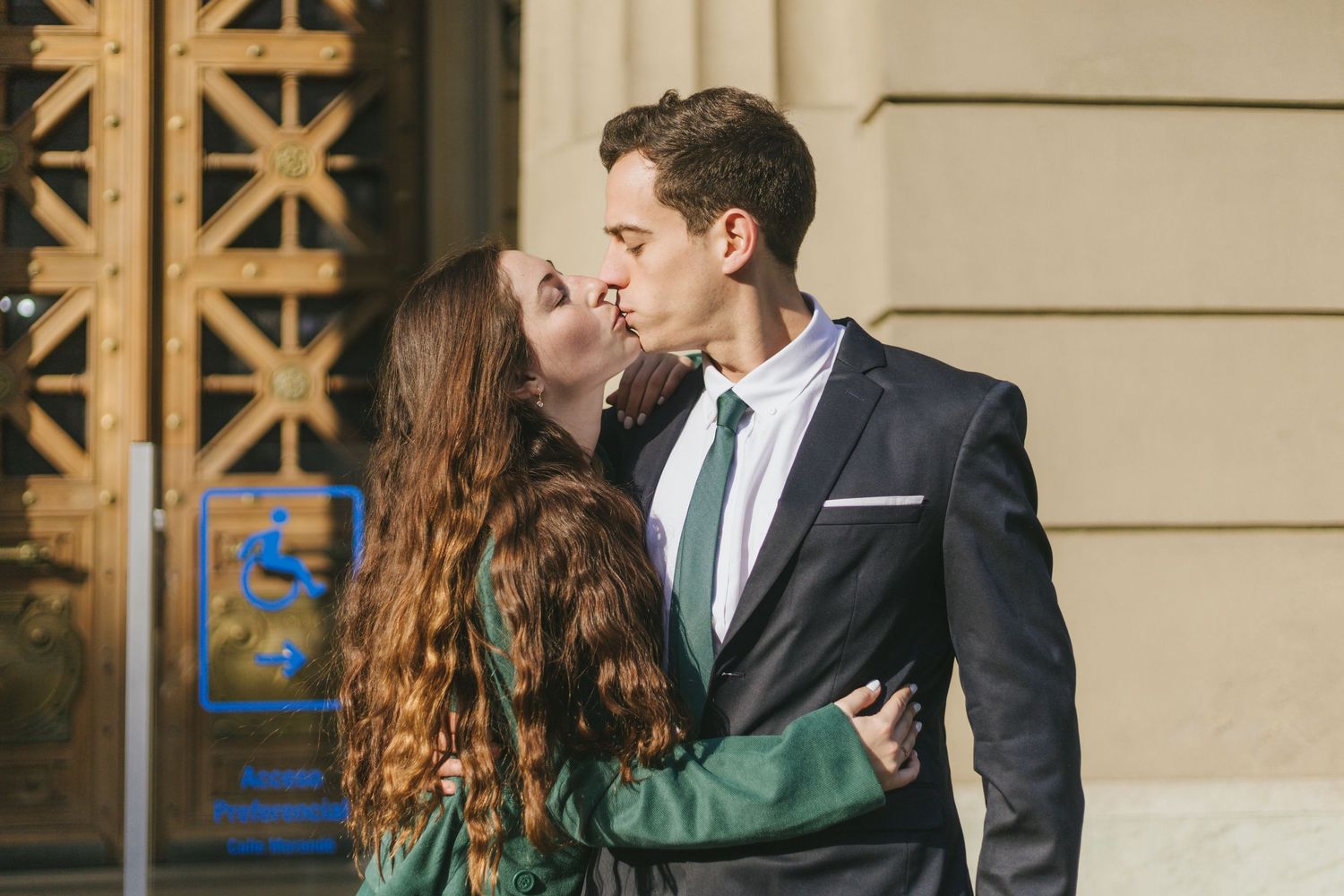 Couple sharing a kiss outside a classical building, woman in green blazer and man in dark suit with green tie.