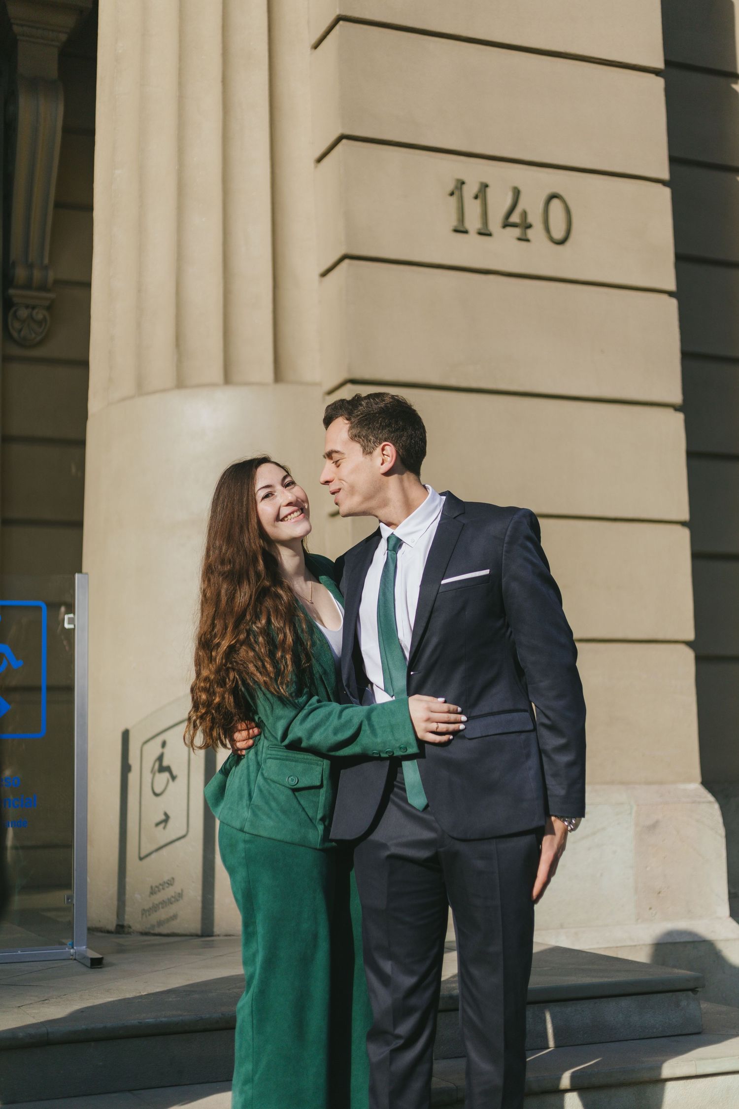 Couple standing close and smiling near a building corner marked 1140, woman in green suit and man in dark suit with green tie.