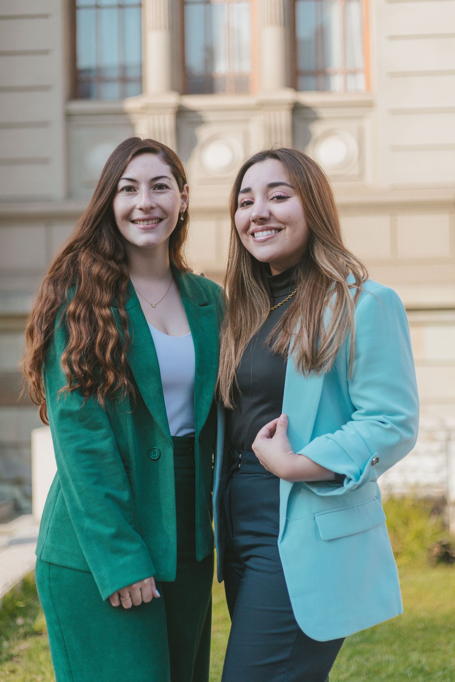 Two smiling women posing outdoors in coordinating blazers, one in emerald green and one in light teal, against a building backdrop.