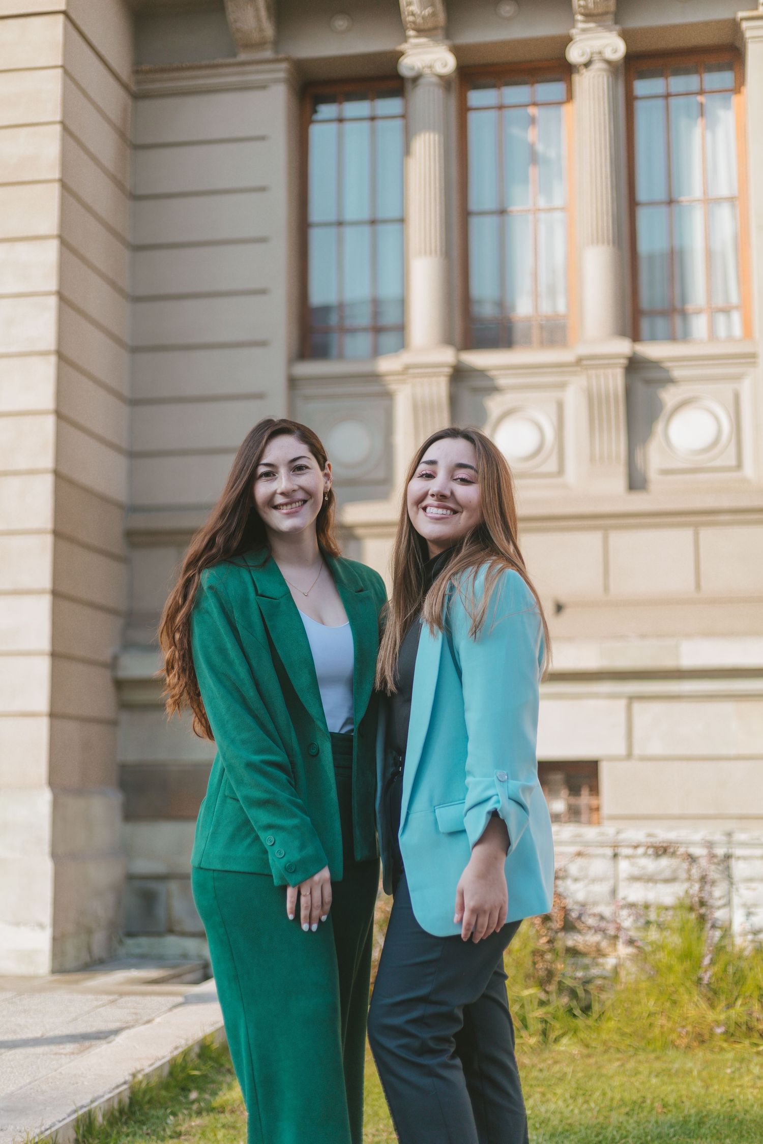 Two cheerful women posing outdoors in matching blazers, one in emerald green and one in teal, with a classical building behind them.