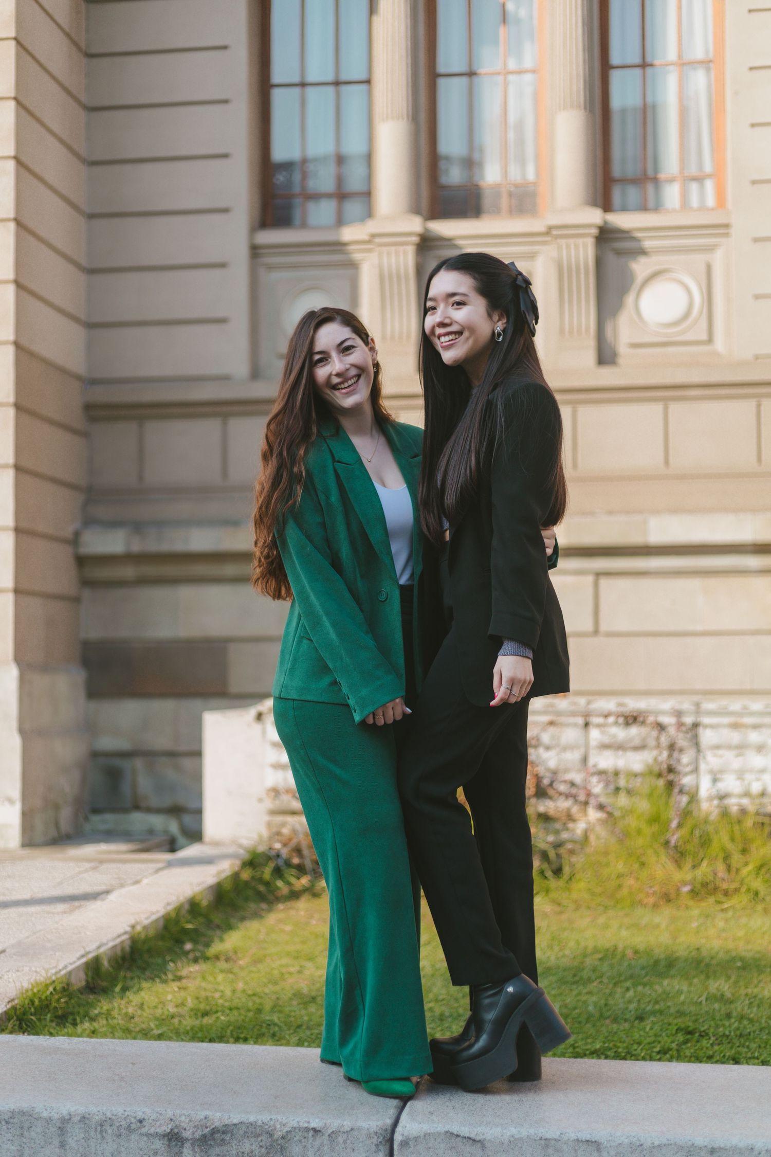 Two young women laughing and holding hands outdoors, one in a green suit and one in a black suit, near a classical building.