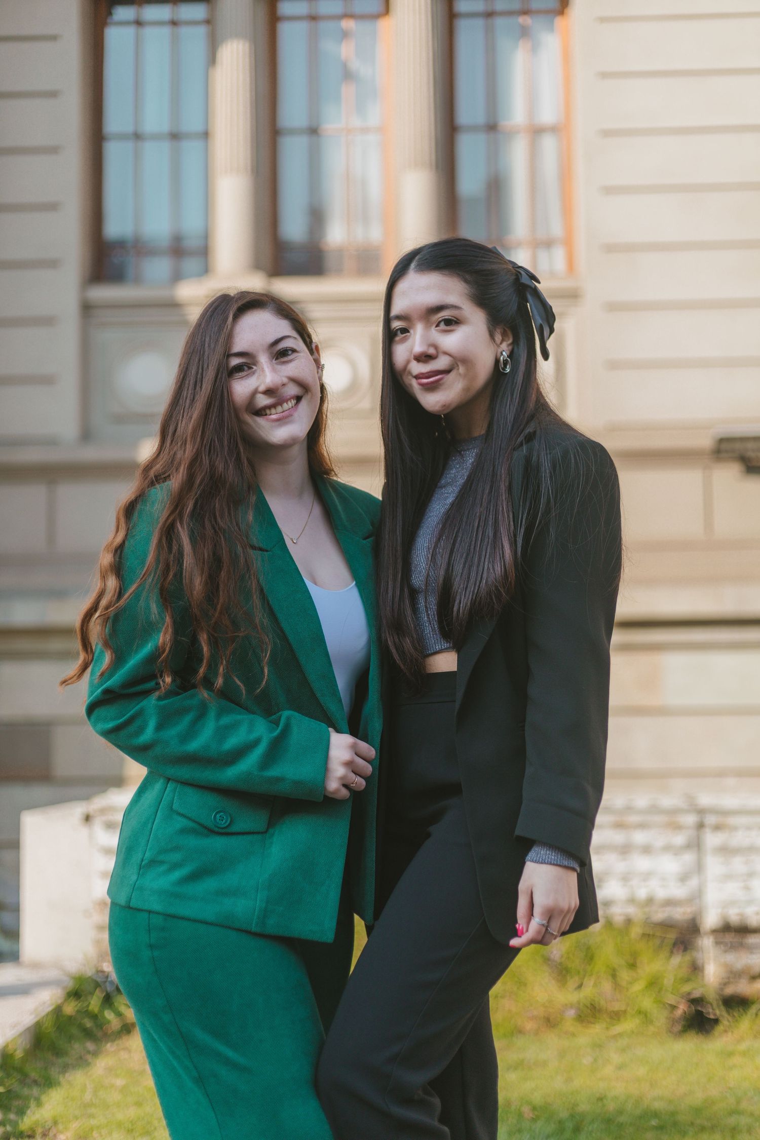 Two smiling women standing together outside a classical building, one in a green suit and one in a black blazer with black pants.