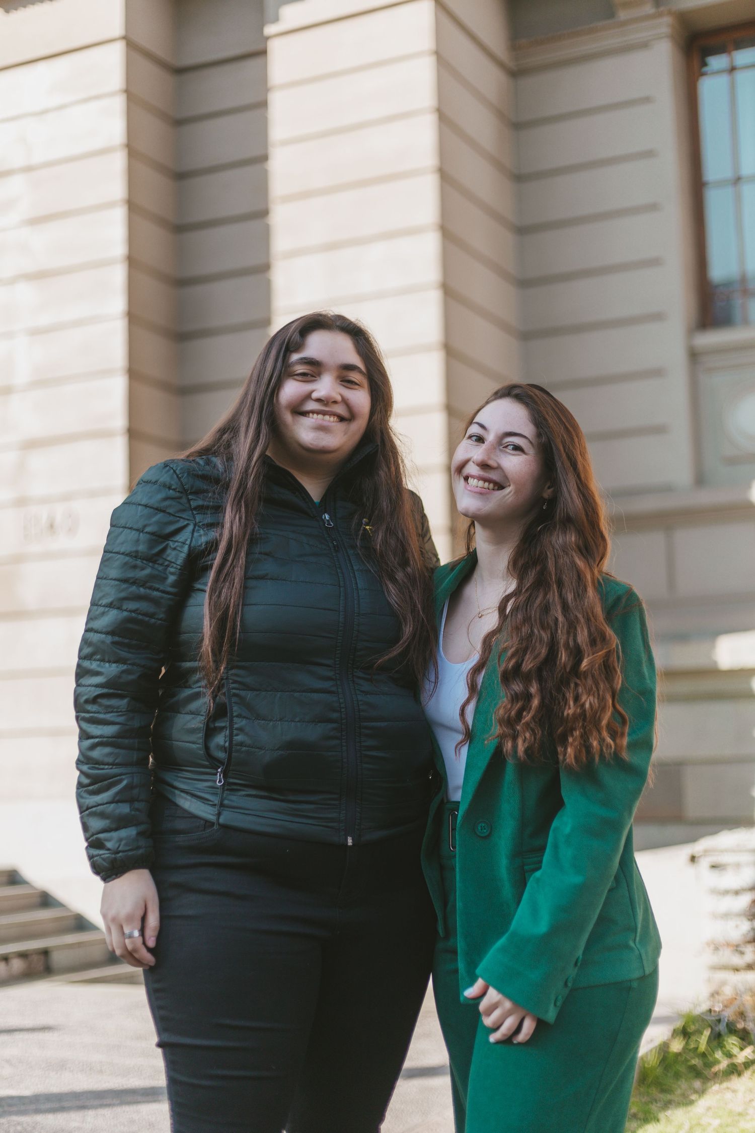 Two young women smiling together outside a stone building, one in a black puffer jacket and one in a green blazer suit.