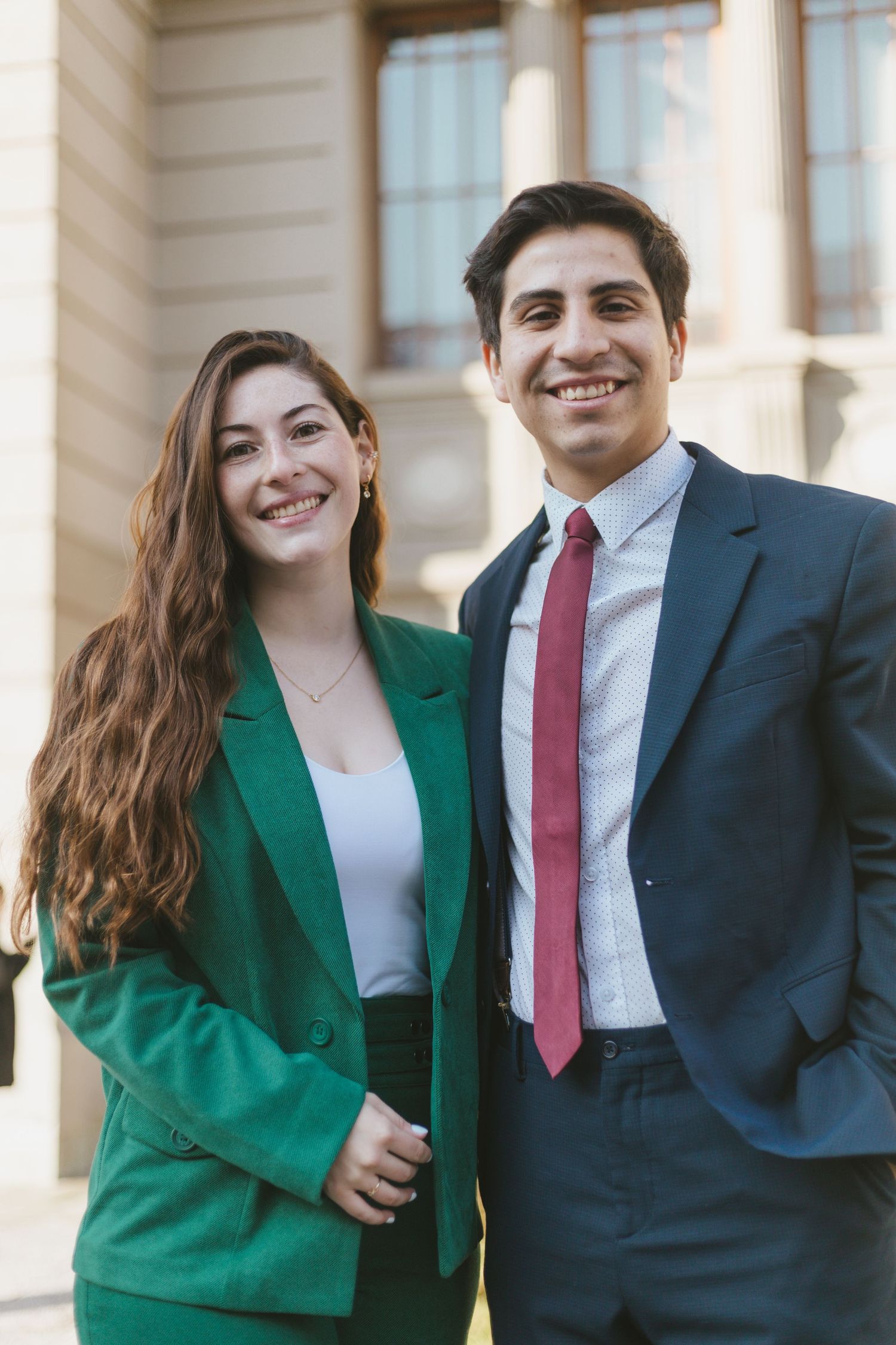 Young man and woman in professional attire smiling outdoors near a classical building.