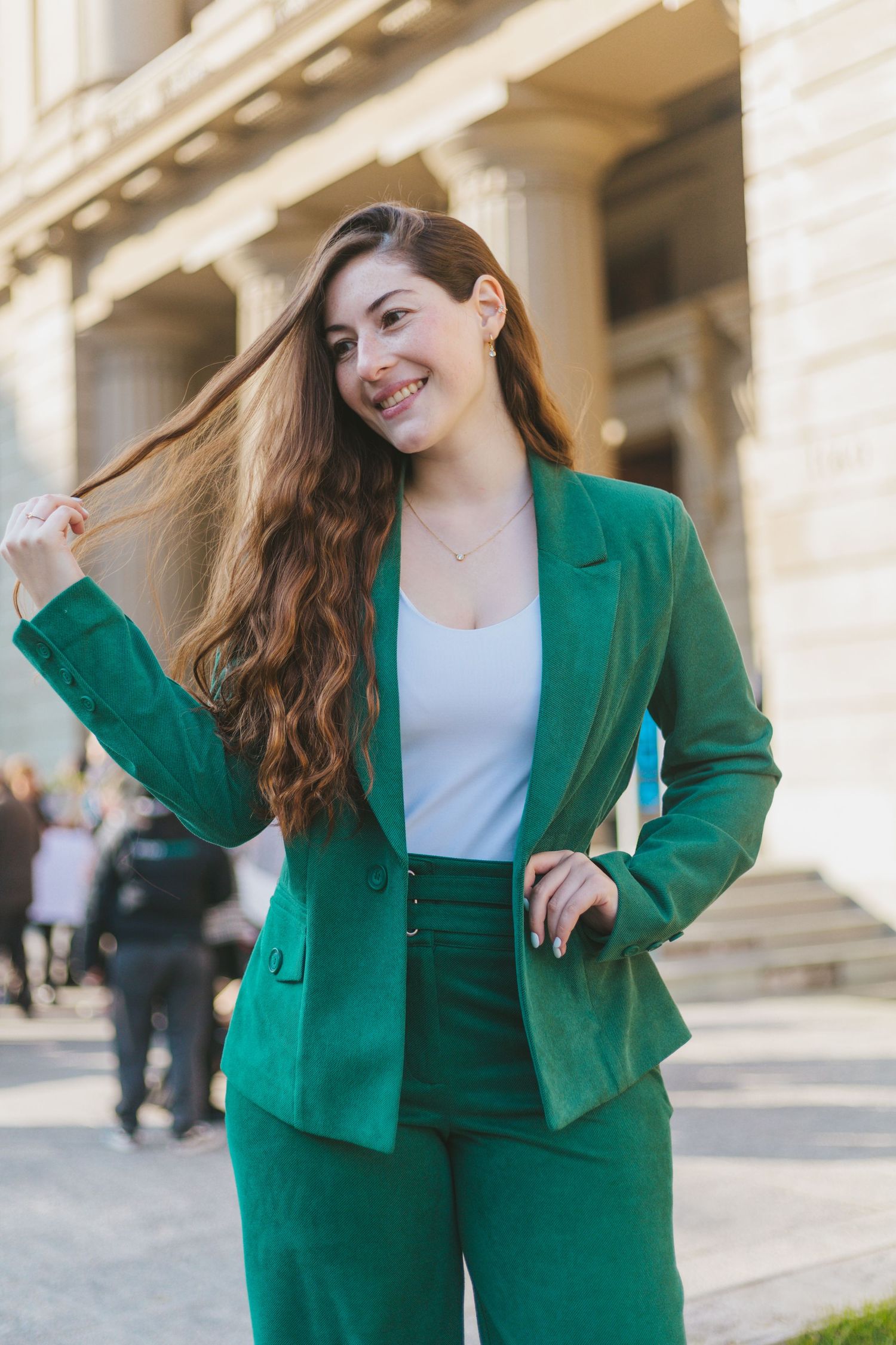 Smiling young woman in green suit touching her hair outdoors near a neoclassical building.