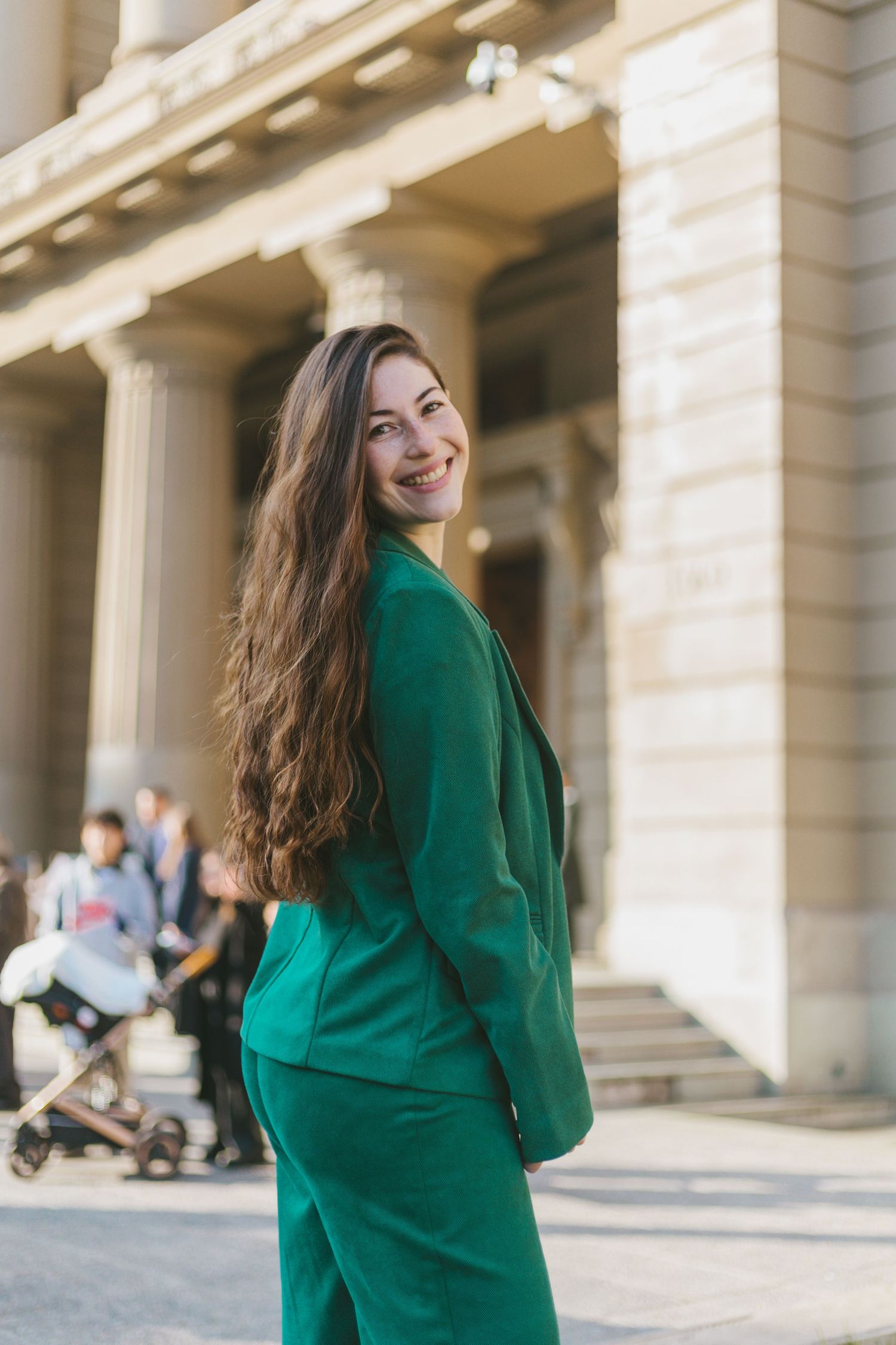 Young woman in green pantsuit smiling over her shoulder outside a classical building.