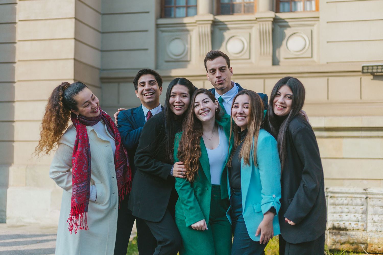 Seven smiling young professionals in formal attire posing together outside a building.