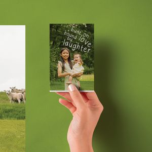 Hand holding a photo card with two women and text built on love and laughter against a green pastoral background.
