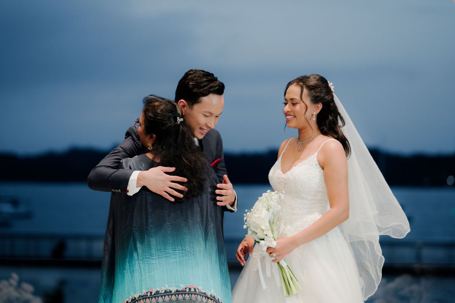 Bride in white gown and groom in suit share a joyful hug with a guest at a waterfront wedding ceremony at dusk.