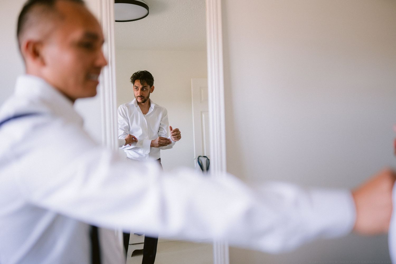 Groom adjusting his white dress shirt while his reflection is visible in a large mirror during wedding preparations.