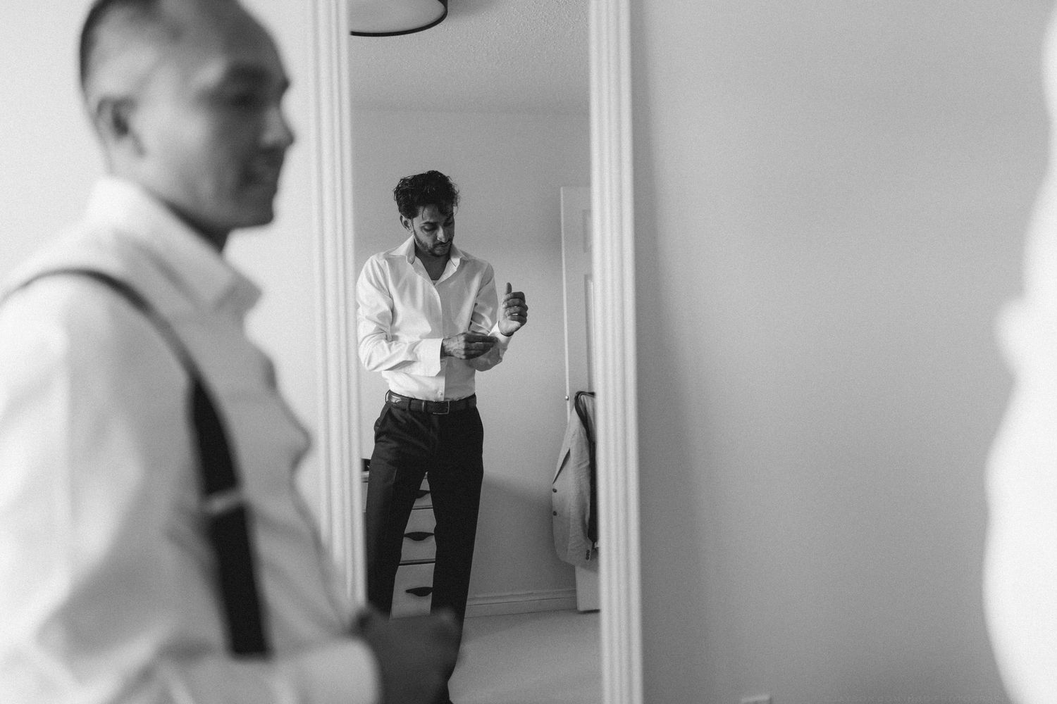 Black and white photo of groom checking his cufflinks in mirror while groomsman stands in foreground.