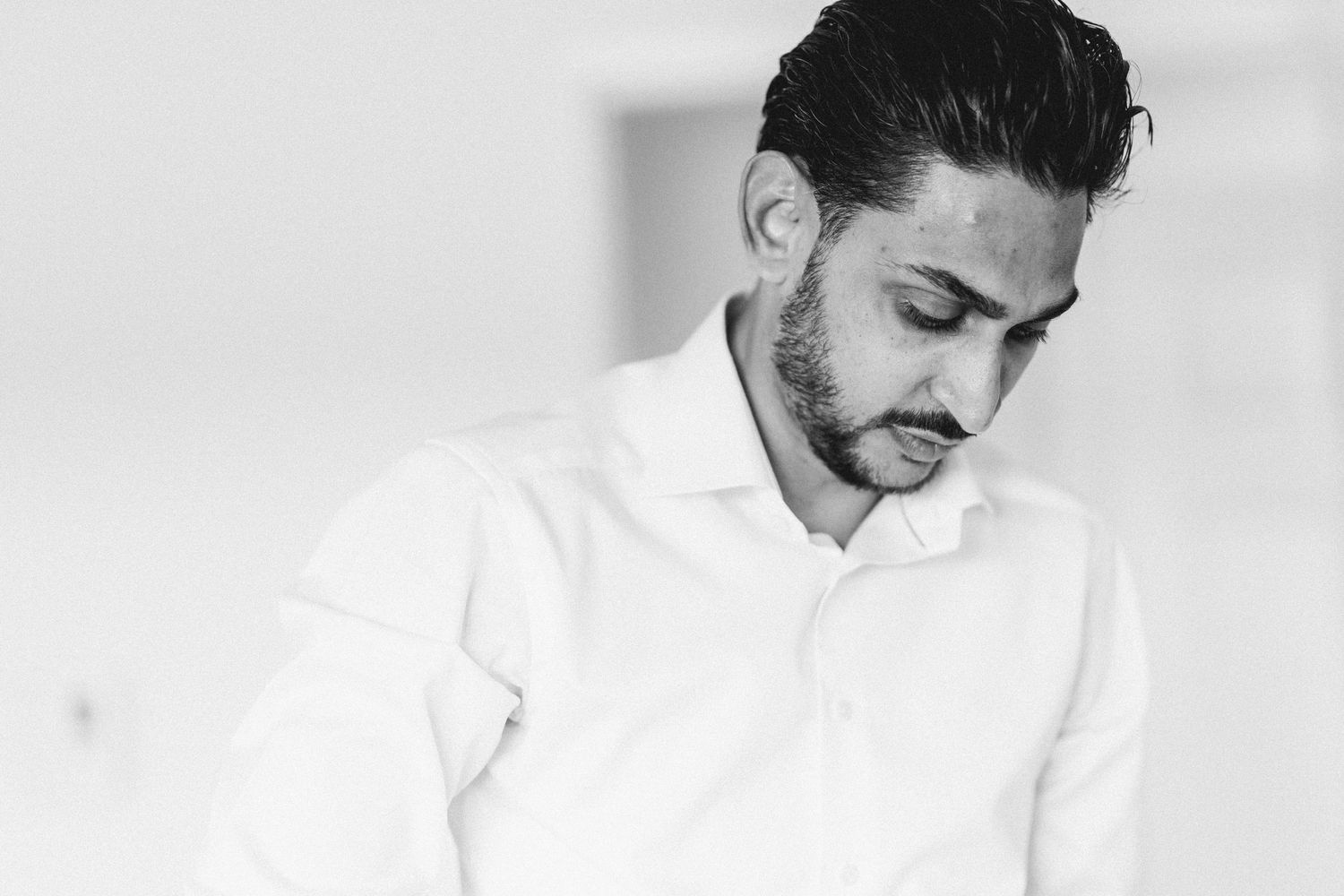 Black and white portrait of groom in white shirt looking down during wedding preparations.