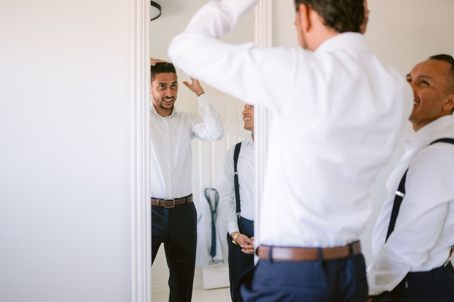 Groom checks his appearance in a mirror while groomsmen assist him in getting dressed for the wedding.