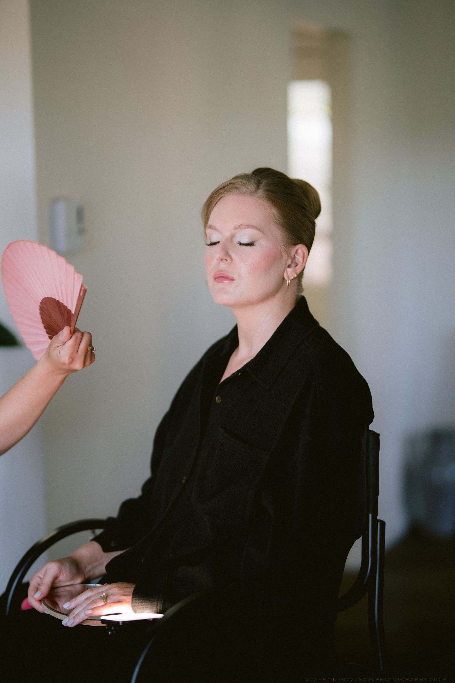 Bride in black coat getting makeup applied while holding a pink fan mirror in a bright room.