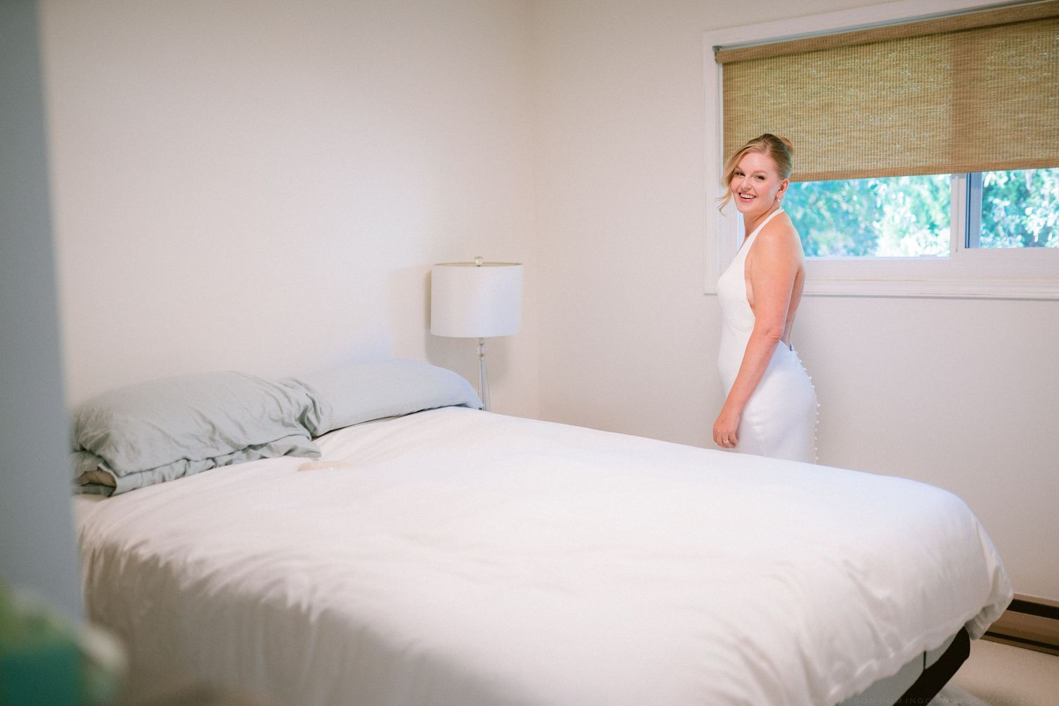 Bride in white wedding dress stands beside bed in bright minimalist bedroom before ceremony.