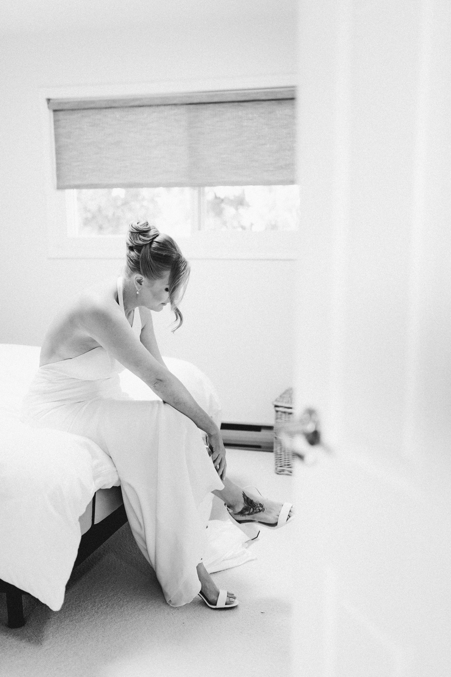 Black and white photo of bride in white halter gown sitting on bed adjusting her shoes.