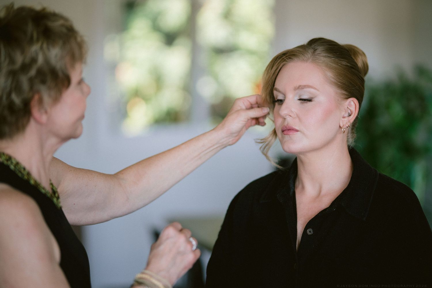 Makeup artist applying finishing touches to blonde bride's face before wedding ceremony indoors.