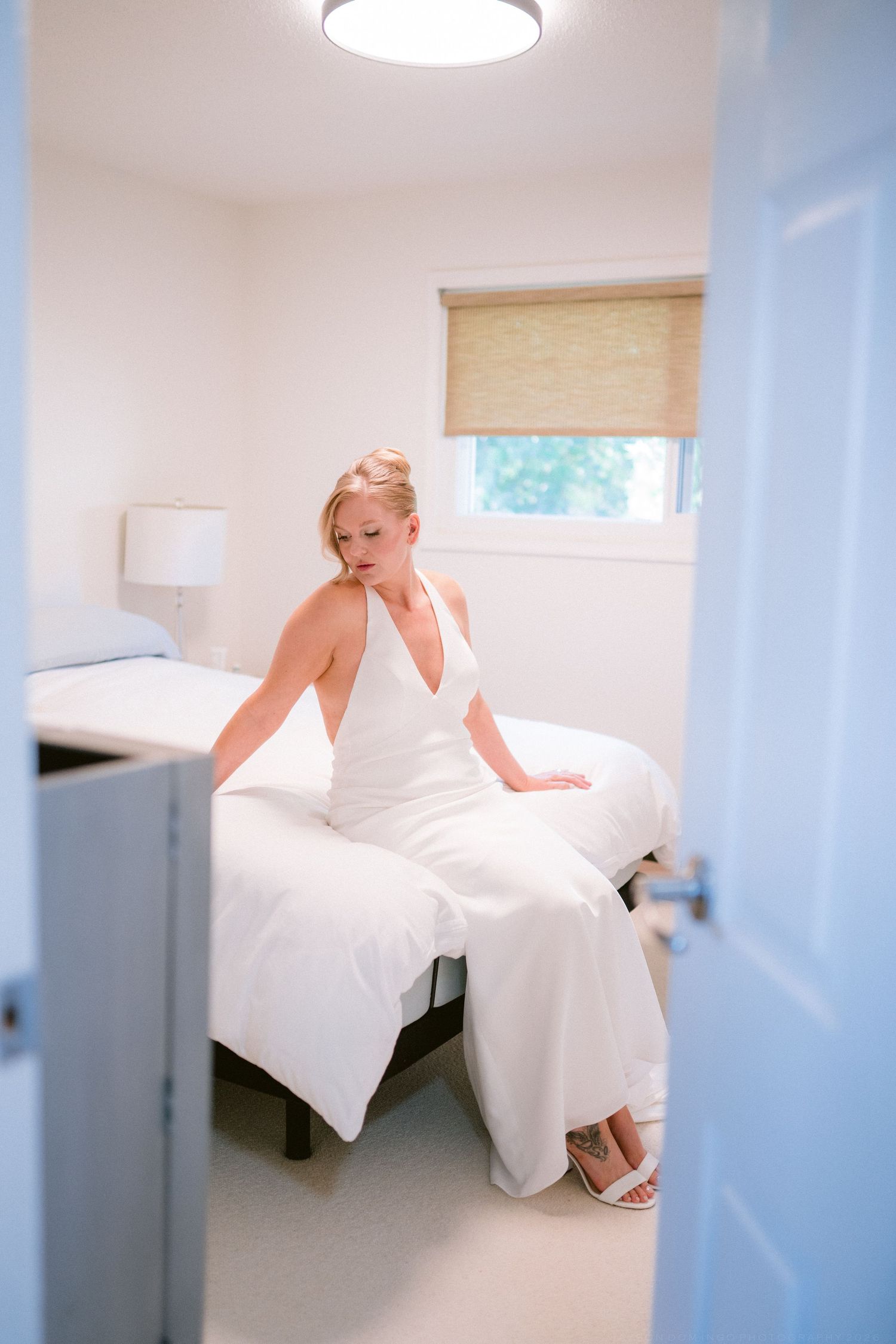 Bride in white halter wedding gown sitting on bed in bright room preparing for wedding day.