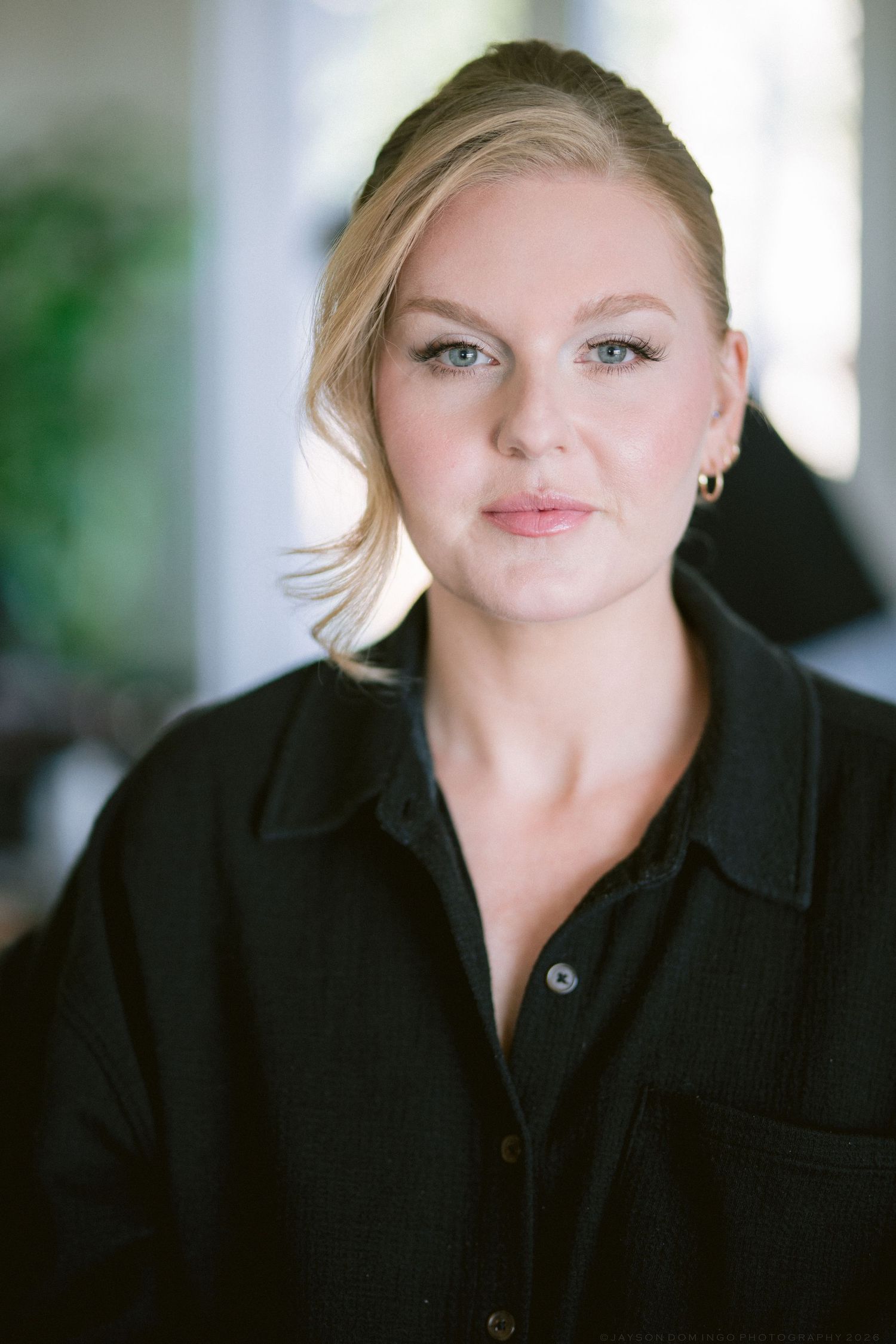 Portrait of blonde woman in black shirt with subtle makeup and gold earrings in soft natural light.