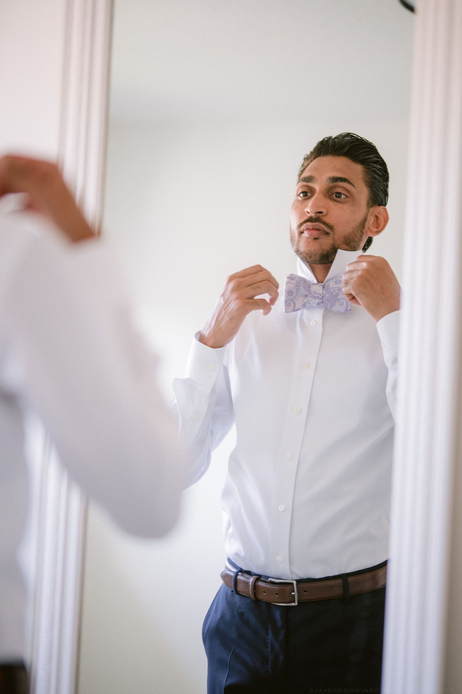 Man adjusting purple bow tie in mirror while wearing white dress shirt and navy pants for formal occasion.
