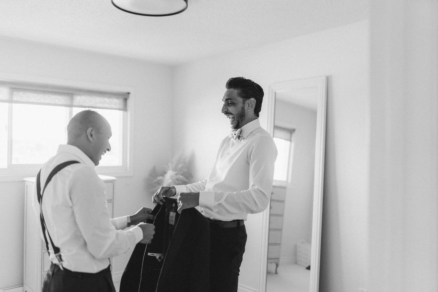 Black and white photo of groom and groomsman laughing while getting ready in bright room.